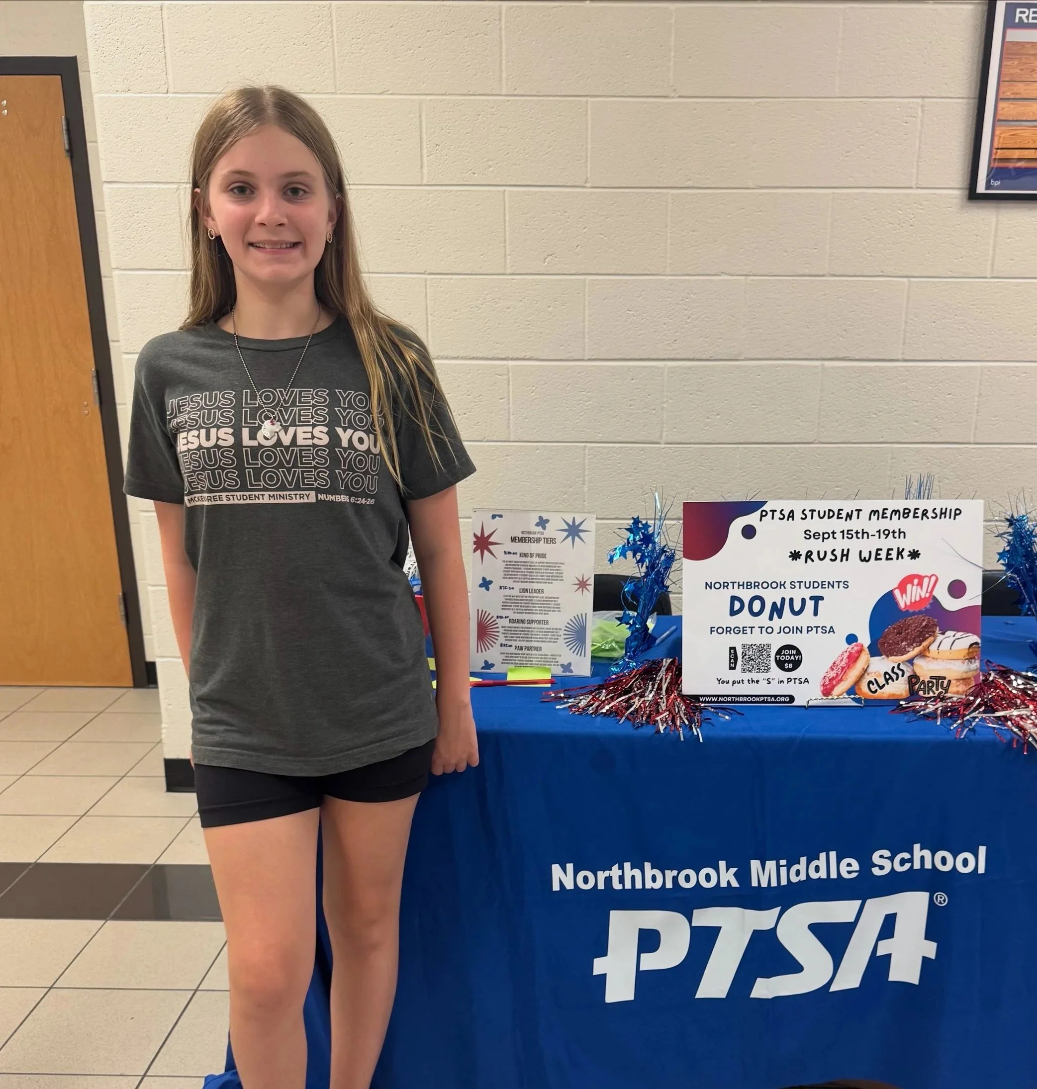 A girl standing next to a display board for Northbrook Middle School PTSA, wearing a black t-shirt with white text and smiling at the camera, inside a school building with beige brick walls and tiled floor.