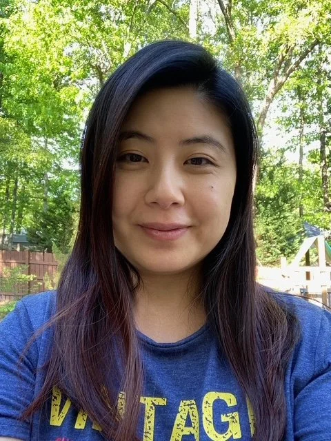 A woman with long dark hair and a slight smile stands outdoors surrounded by green trees, wearing a blue shirt.