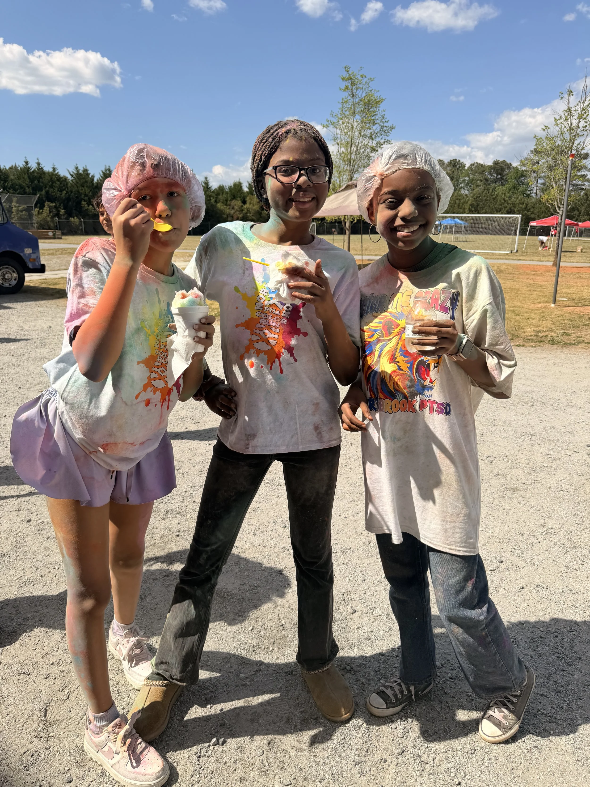 Three young girls covered in colorful powder enjoying a festival outdoors on a sunny day, with trees, tents, and a soccer goal in the background.