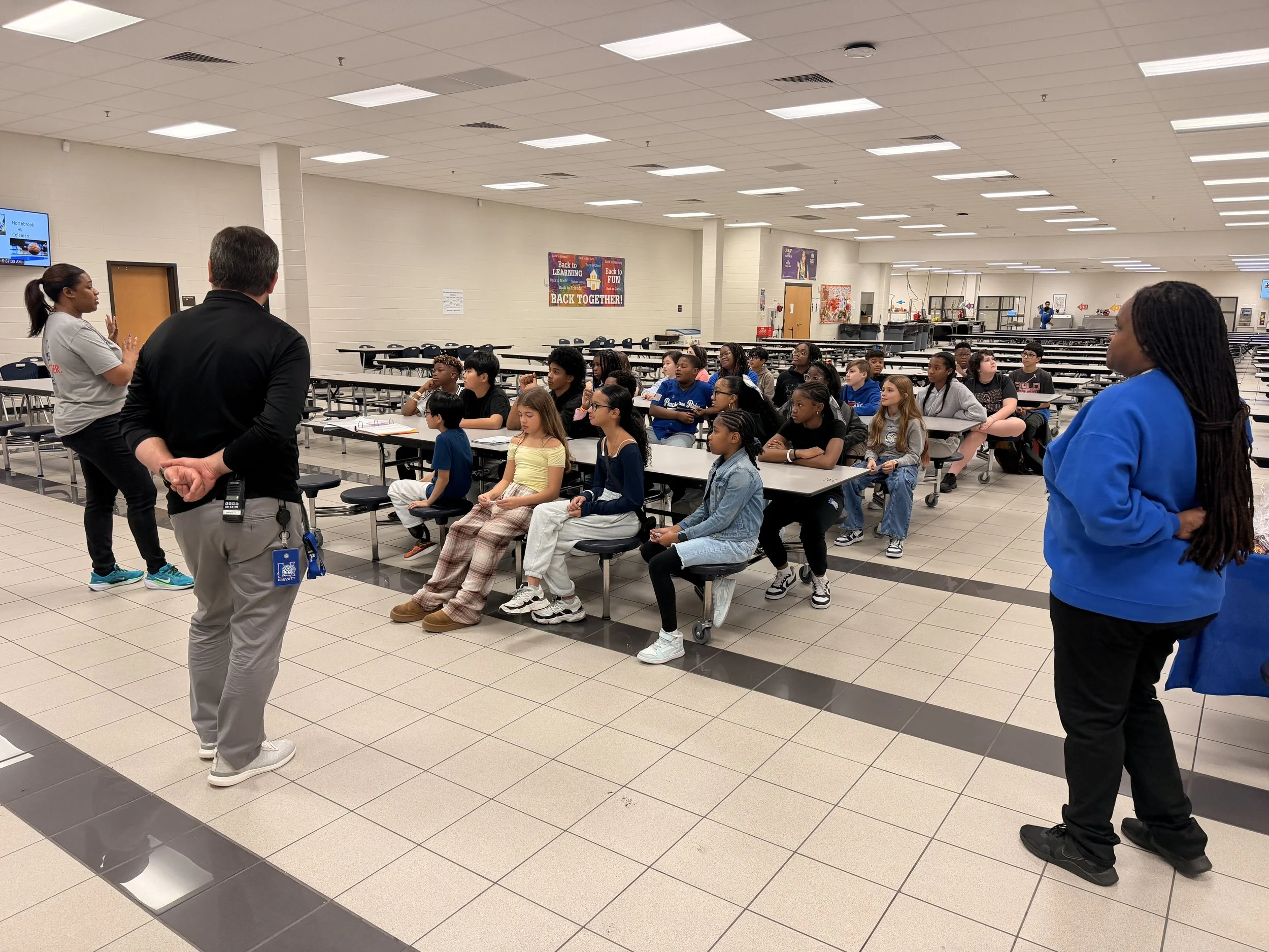 A group of children sitting at cafeteria tables listening to two adults standing and speaking to them in a school cafeteria.