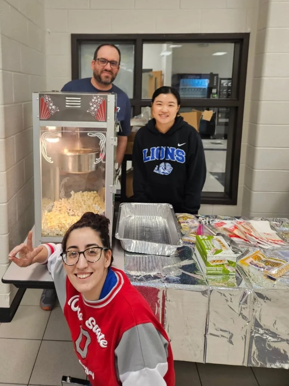 Four people posing around a popcorn machine and a table with snacks in a room with beige brick walls and a windowed room in the background.