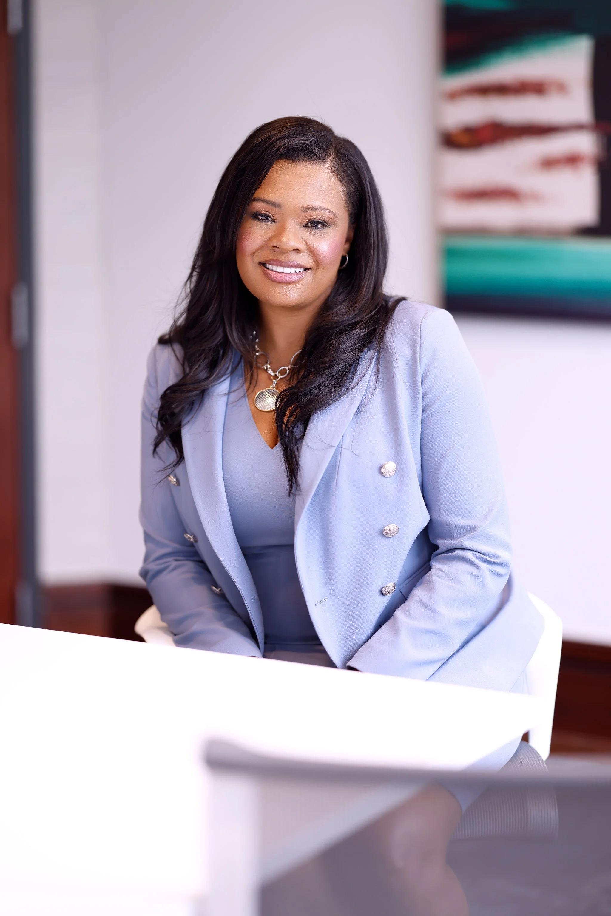 A smiling woman with long black hair, dressed in a light gray blazer and jewelry, sitting at a table in a modern office.