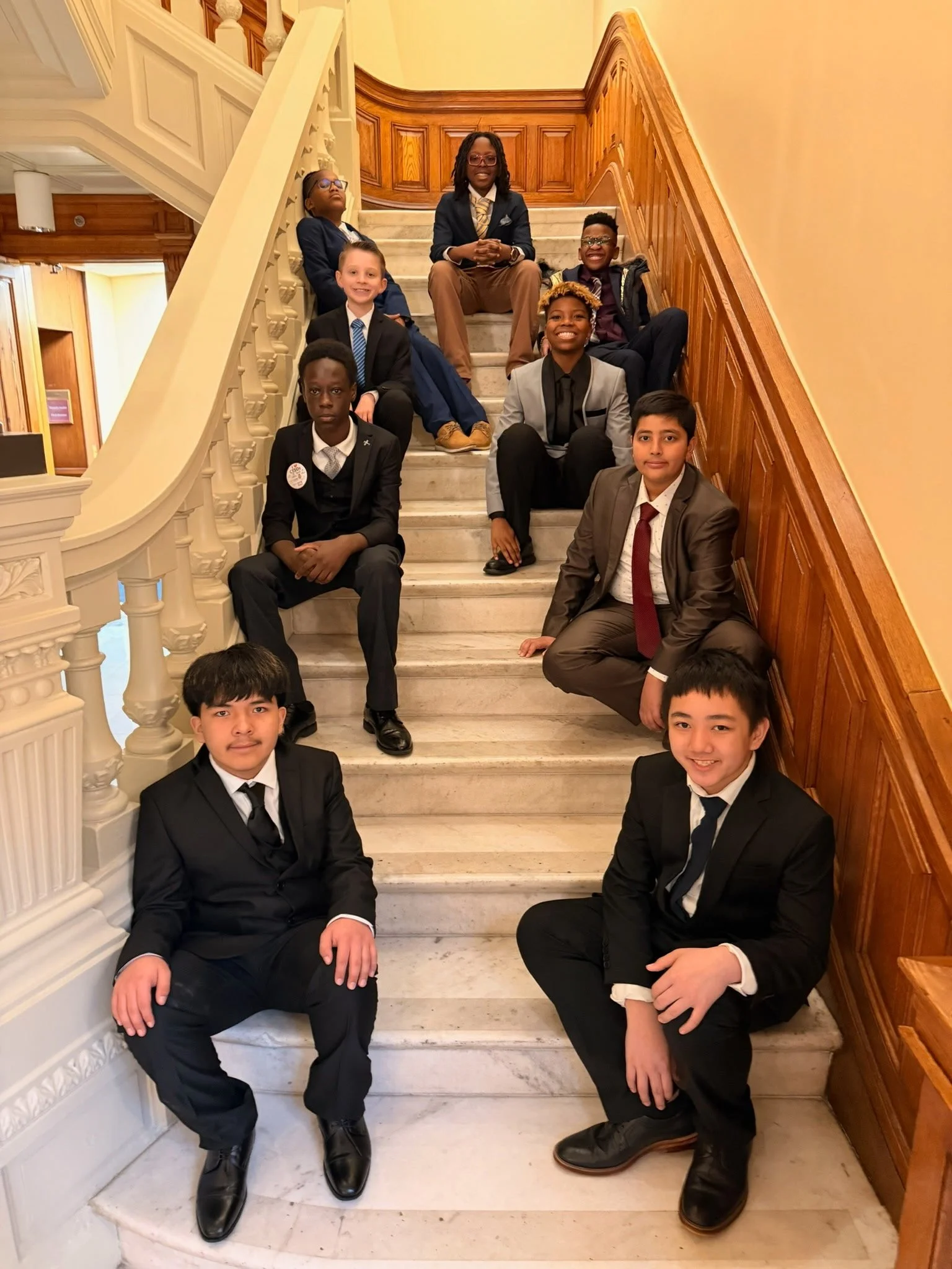 A group of nine young people sitting and standing on a marble staircase in a formal setting, dressed in suits and business attire.