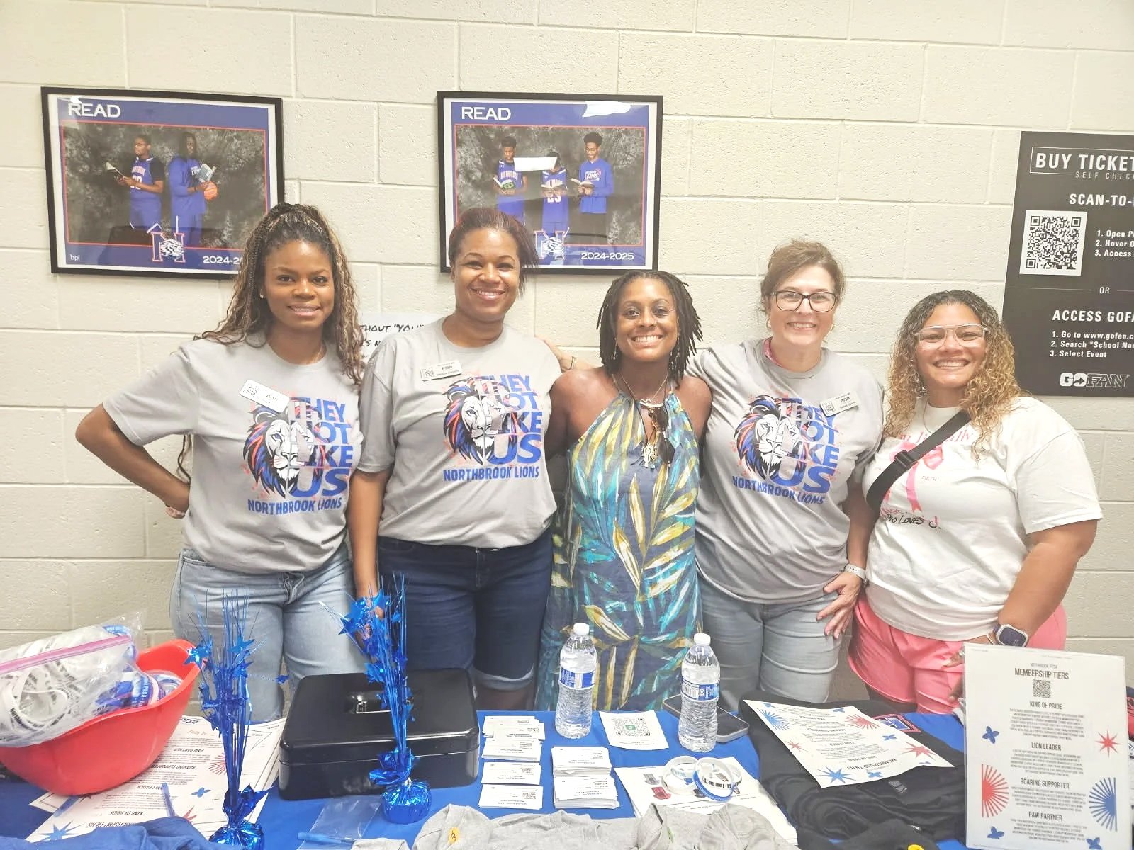 Group of five women standing behind a table with informational materials, wearing t-shirts, in an indoor setting with two framed photos on the wall behind them.
