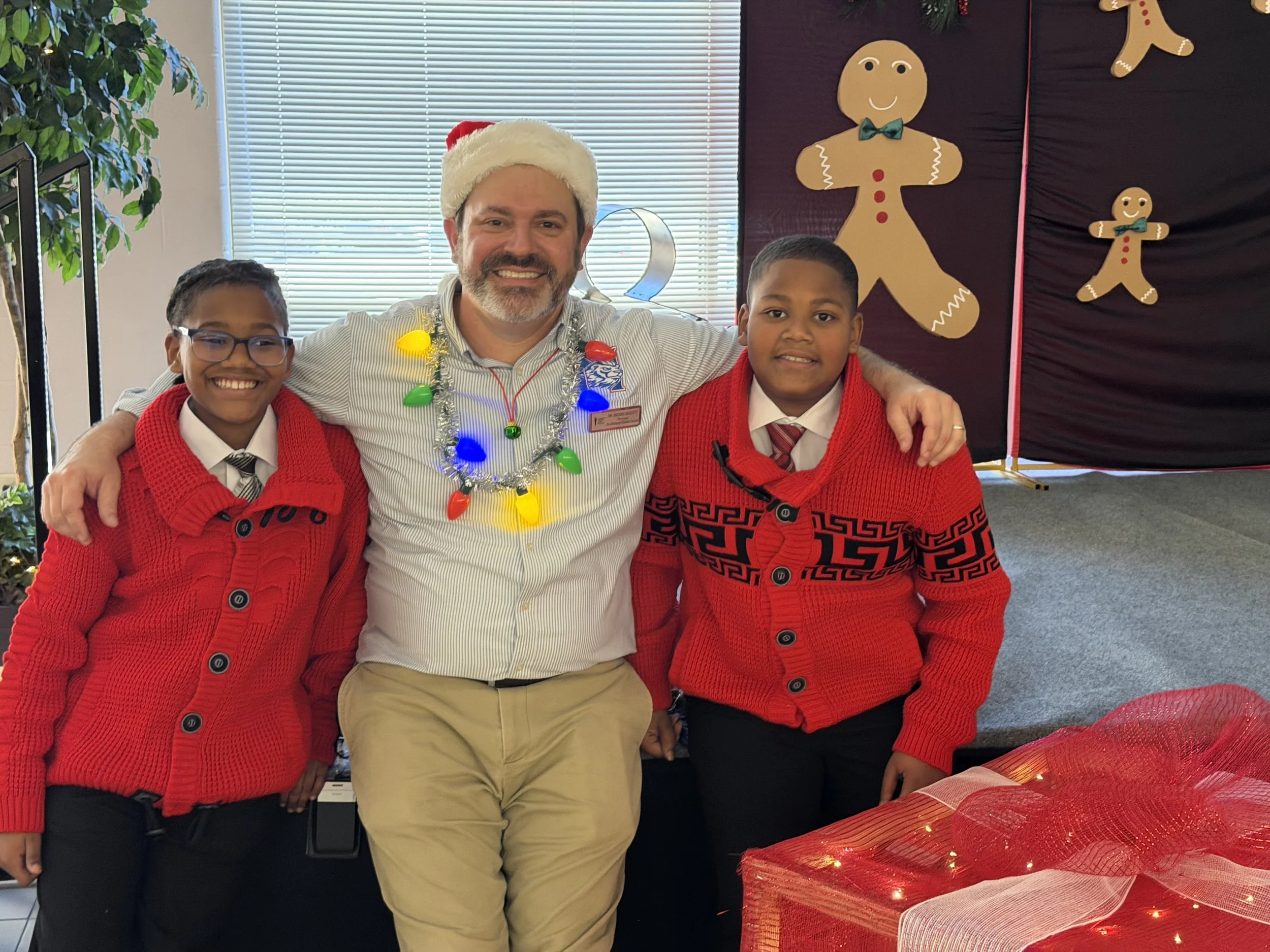 A man wearing a Santa hat and colorful holiday lights around his neck, smiling with two young boys in red sweaters, posed together indoors with a decorated holiday backdrop featuring gingerbread men and a Christmas tree.
