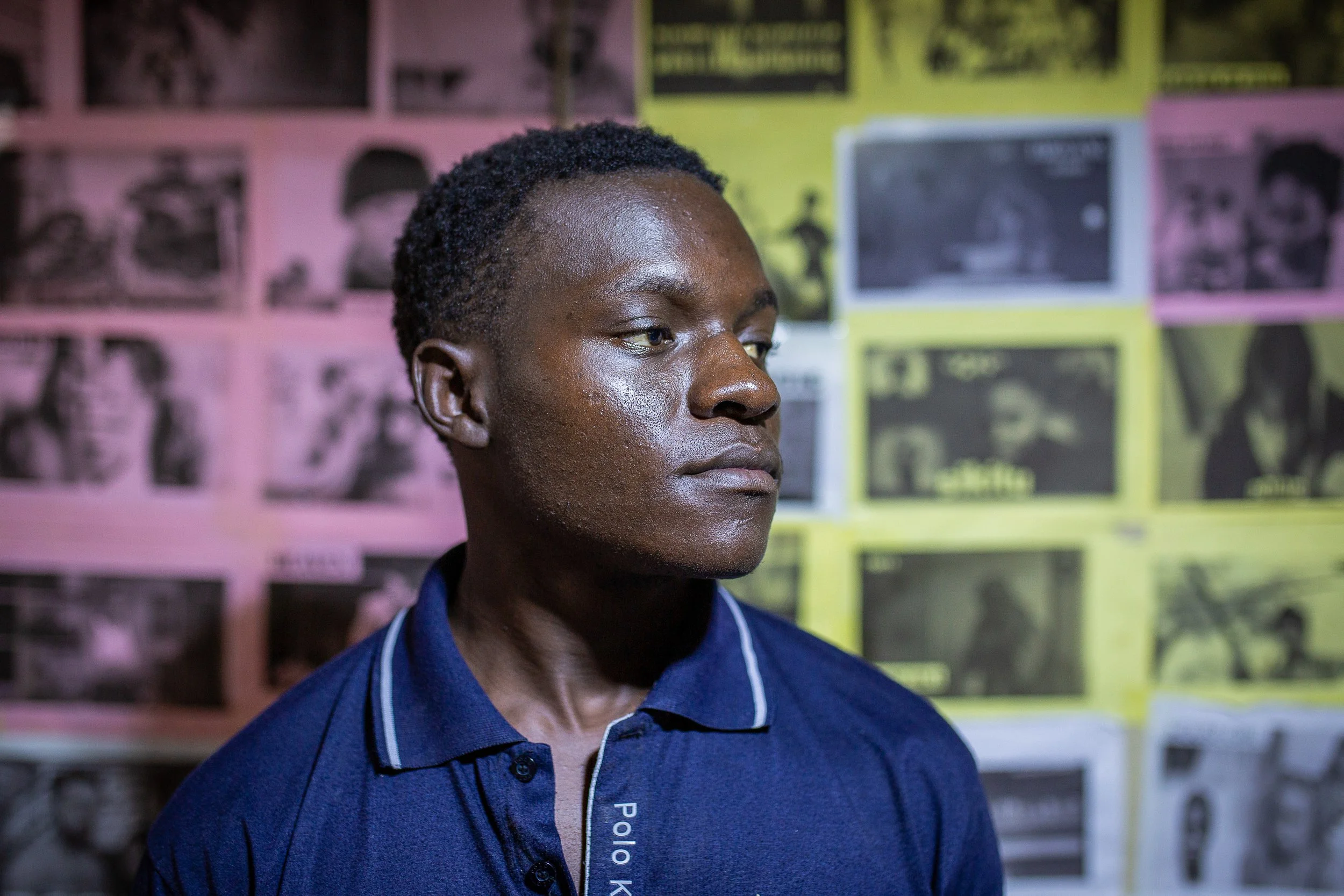Alfred looks off to his left and stands against a wall of black and white photos printed on white, yellow and pink paper.