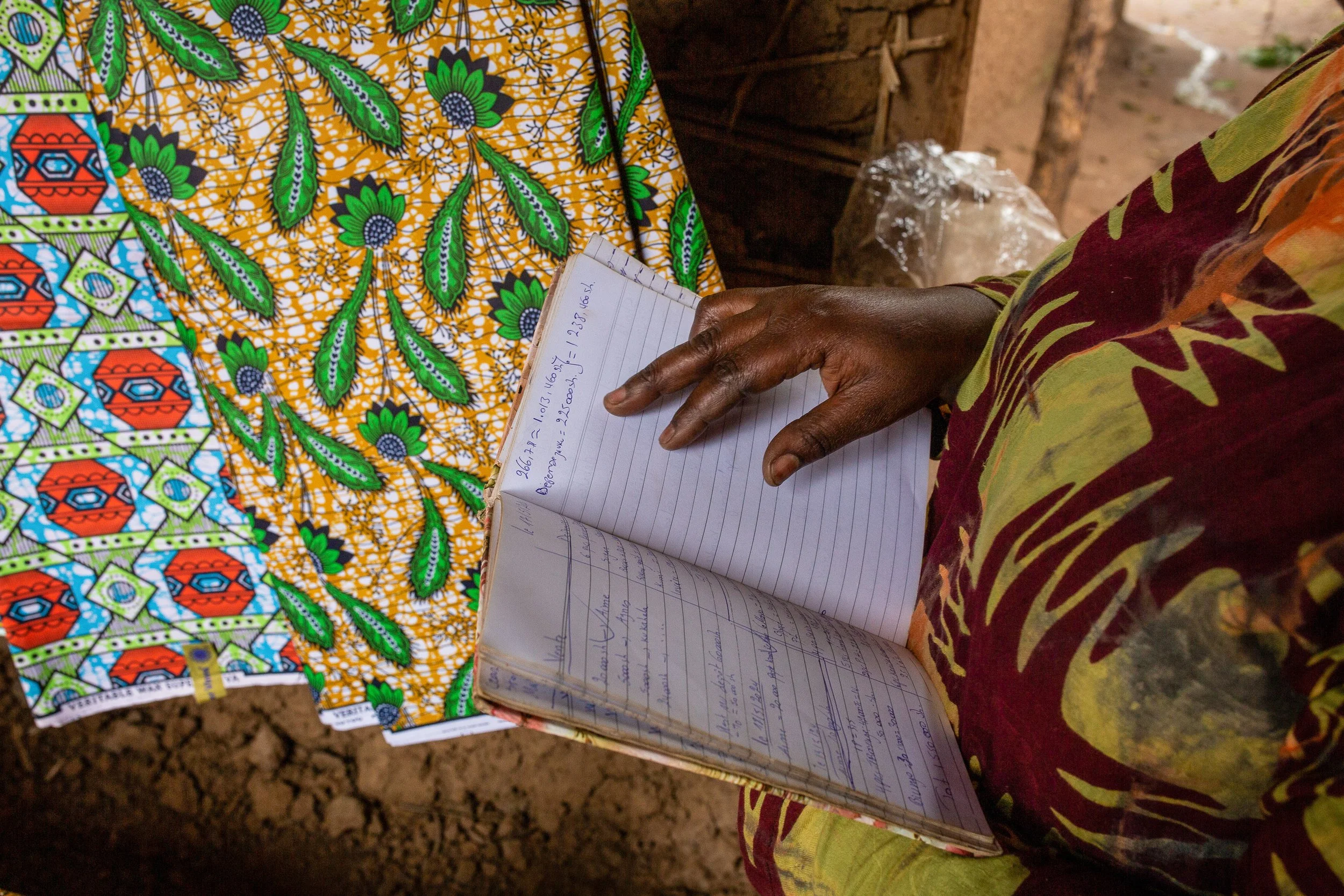 Furaha reads from her bookeeping book against a backdrop of colourful African fabric wax prints. 