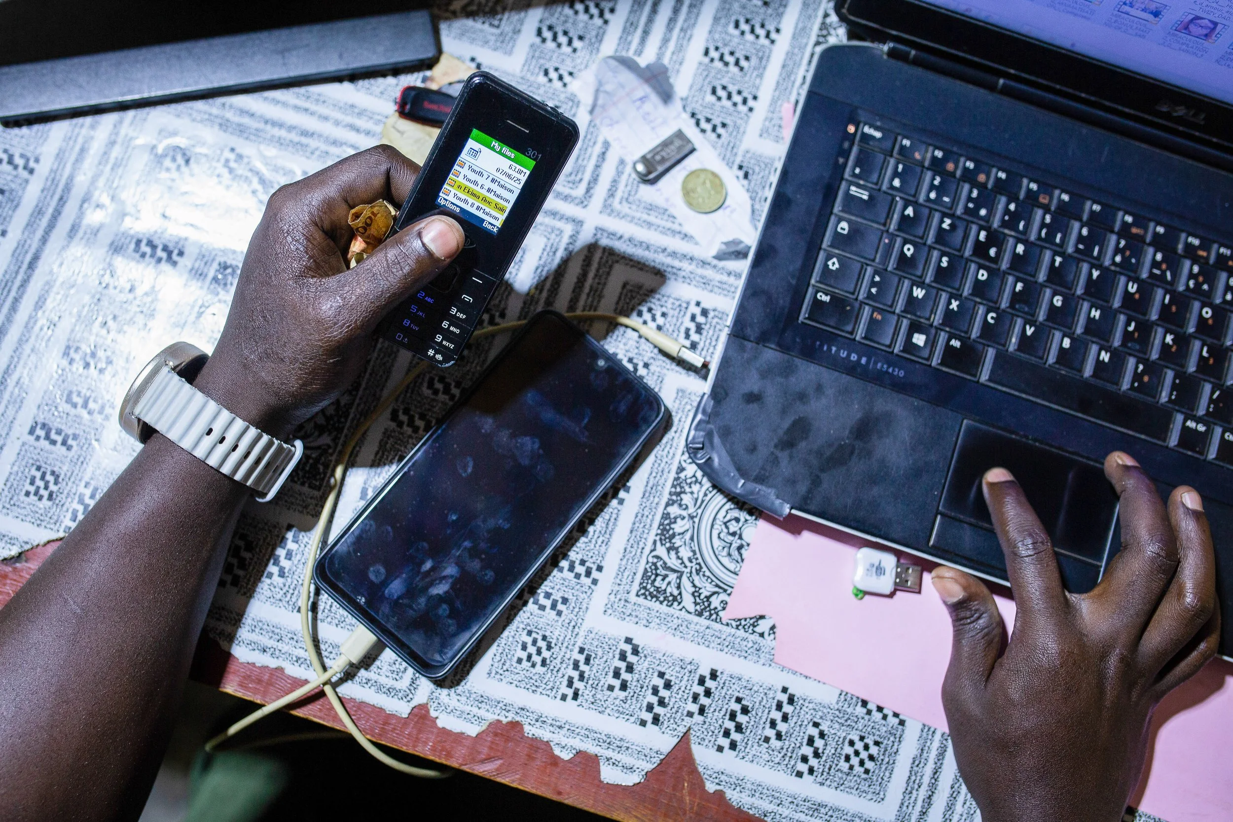 Alfred serves a customer at his stationery store using a basic mobile phone and his laptop.