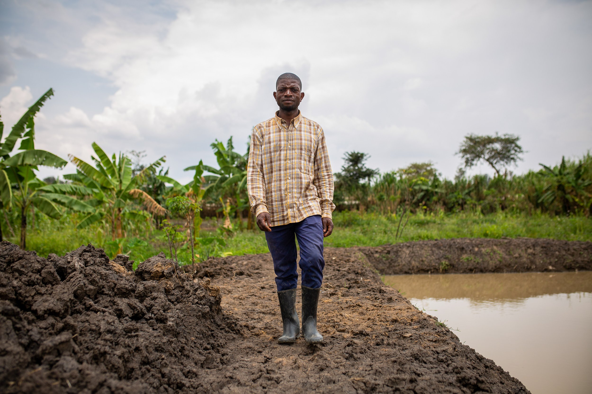Joshua walks next to his fish farm, wearing wellies and a chequered shirt. .