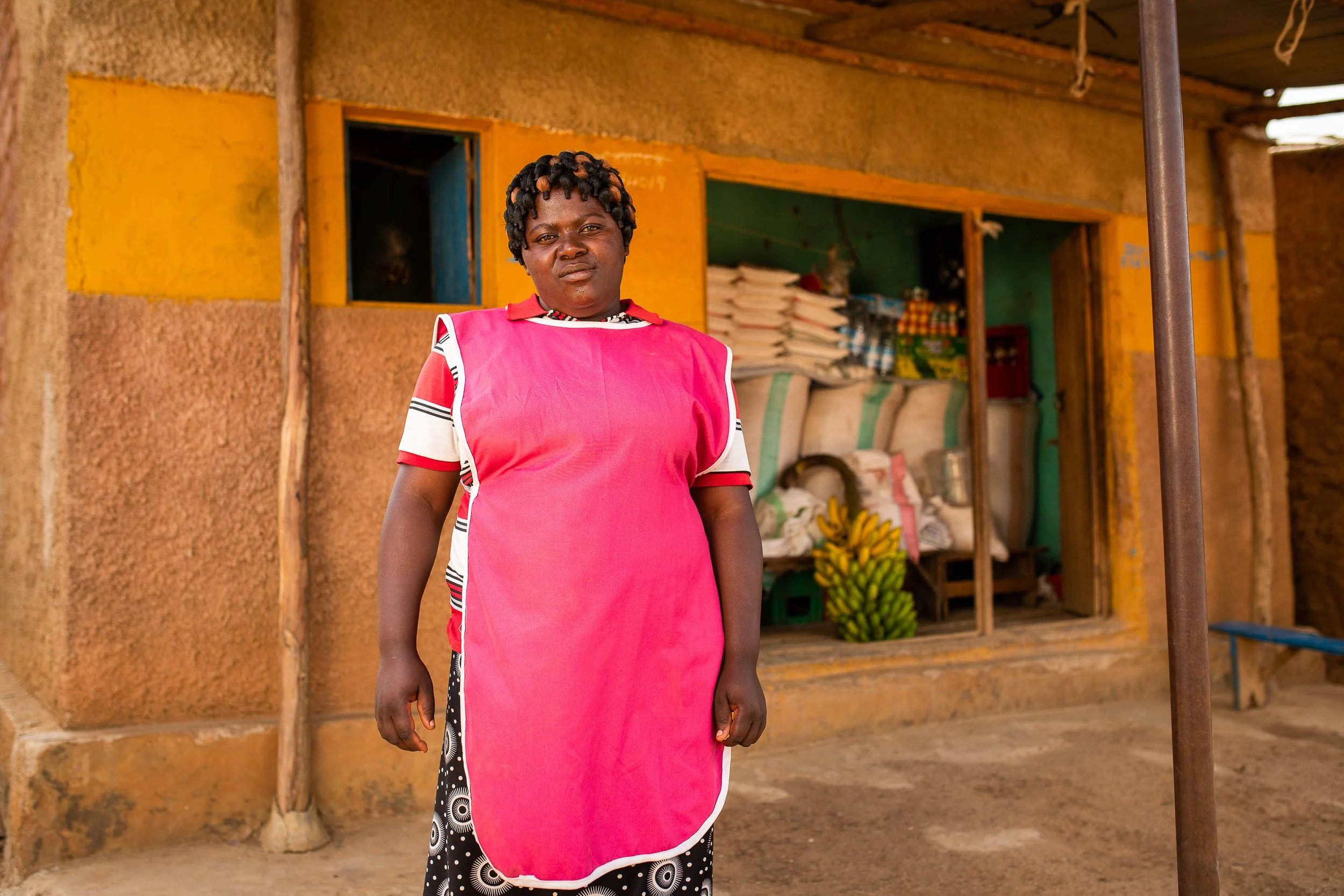 Mediatrice smiles gently as she stands outside her grocery shop. Through her shop door you can see sacks and products piled up and big bunch of bananas.