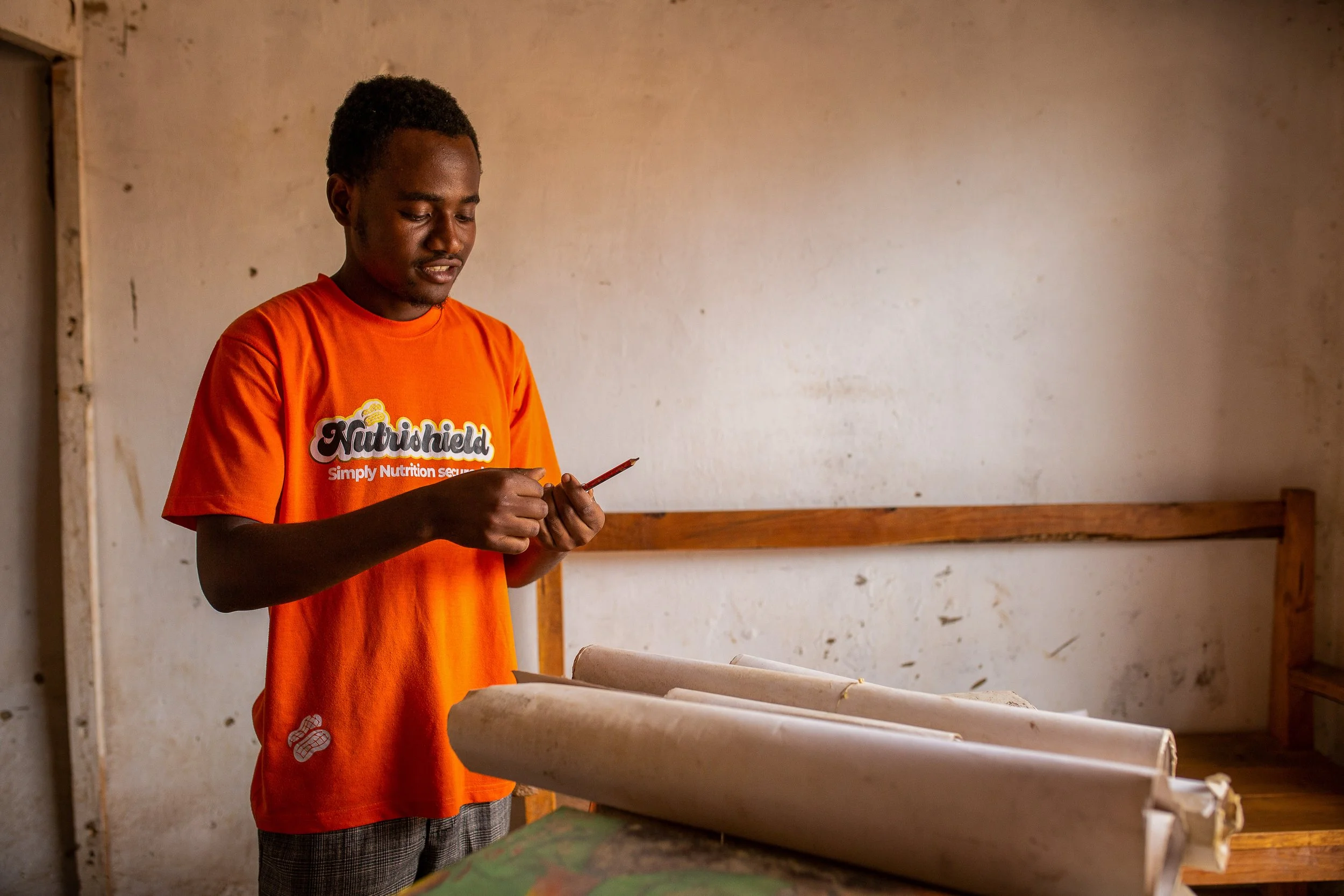 Savino prepares his art supplies. He holds a pencil and a big roll of paper sits on the table in front of him. 