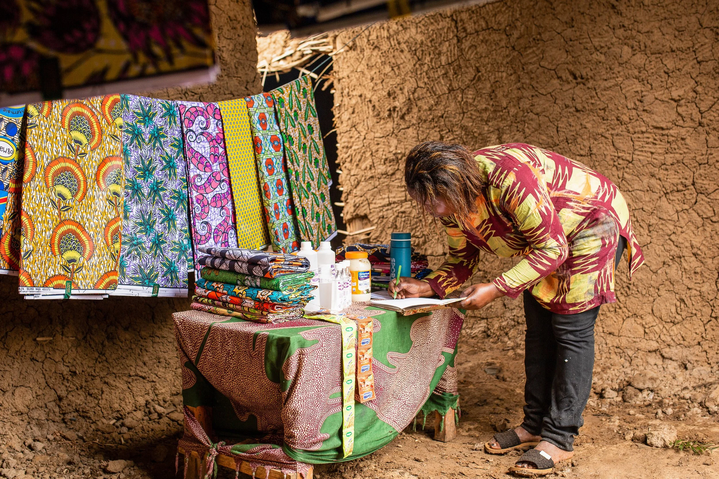 Furaha does the bookkeeping at her fabric shop. She leans down on a table to write in her notebook against a backdrop of colourful fabrics hanging up. 