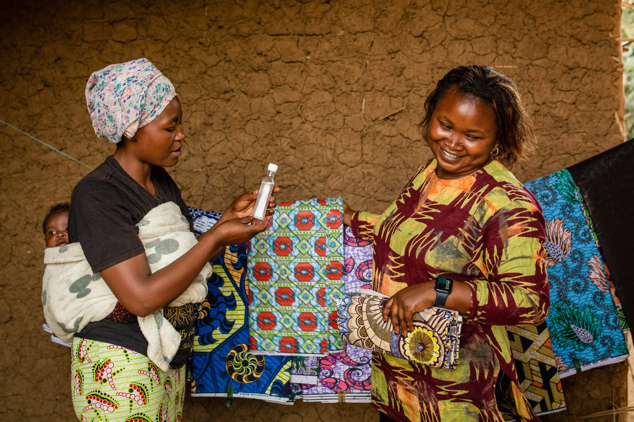 Furaha serves a customer at her shop. They laugh and hold a piece of fabric between them. 