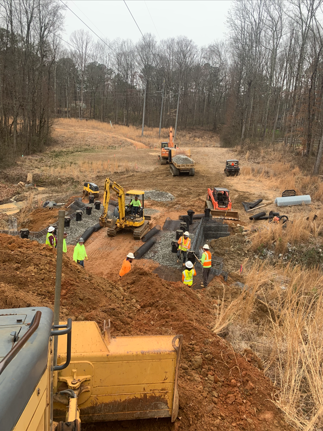 Construction site with workers in safety vests and helmets installing infrastructure, including pipes and a foundation, using construction equipment like excavators and a bulldozer in a rural area with trees and power lines.