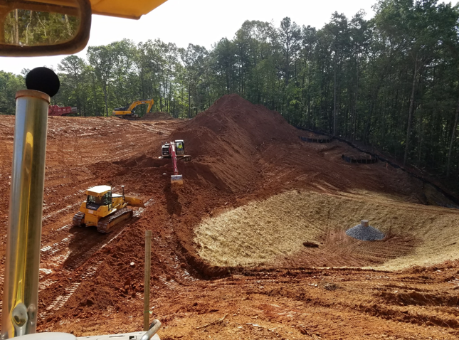 Construction site with heavy machinery on a slope, excavating earth to create a pond or water feature, surrounded by trees.