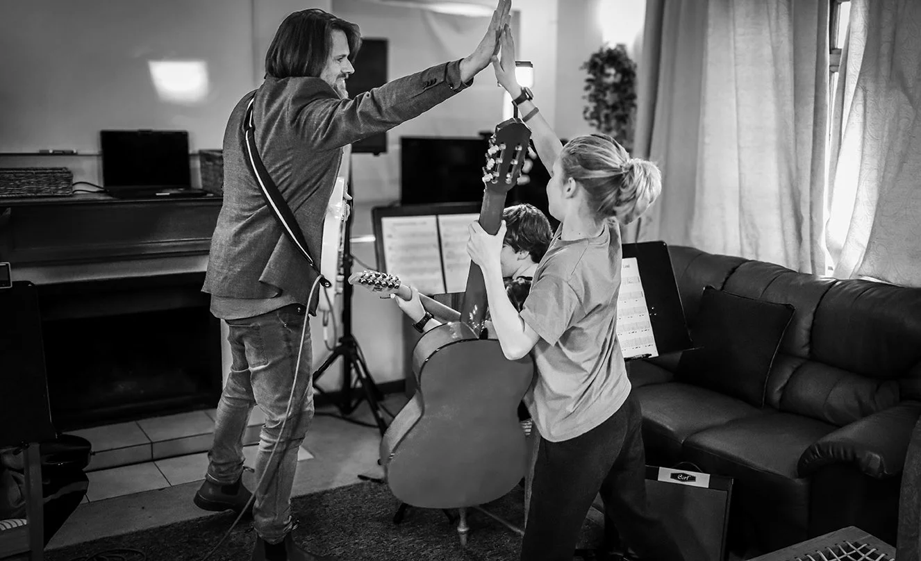 A man and a girl giving each other a high five while holding guitars in a living room with someone playing music in the background.