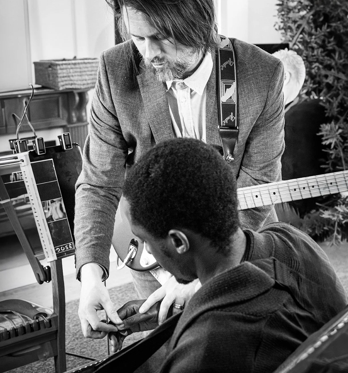 A man in a suit and with a guitar is assisting a person in a wheelchair with a medical procedure or therapy. The scene takes place indoors, with some furniture and a Christmas tree in the background.