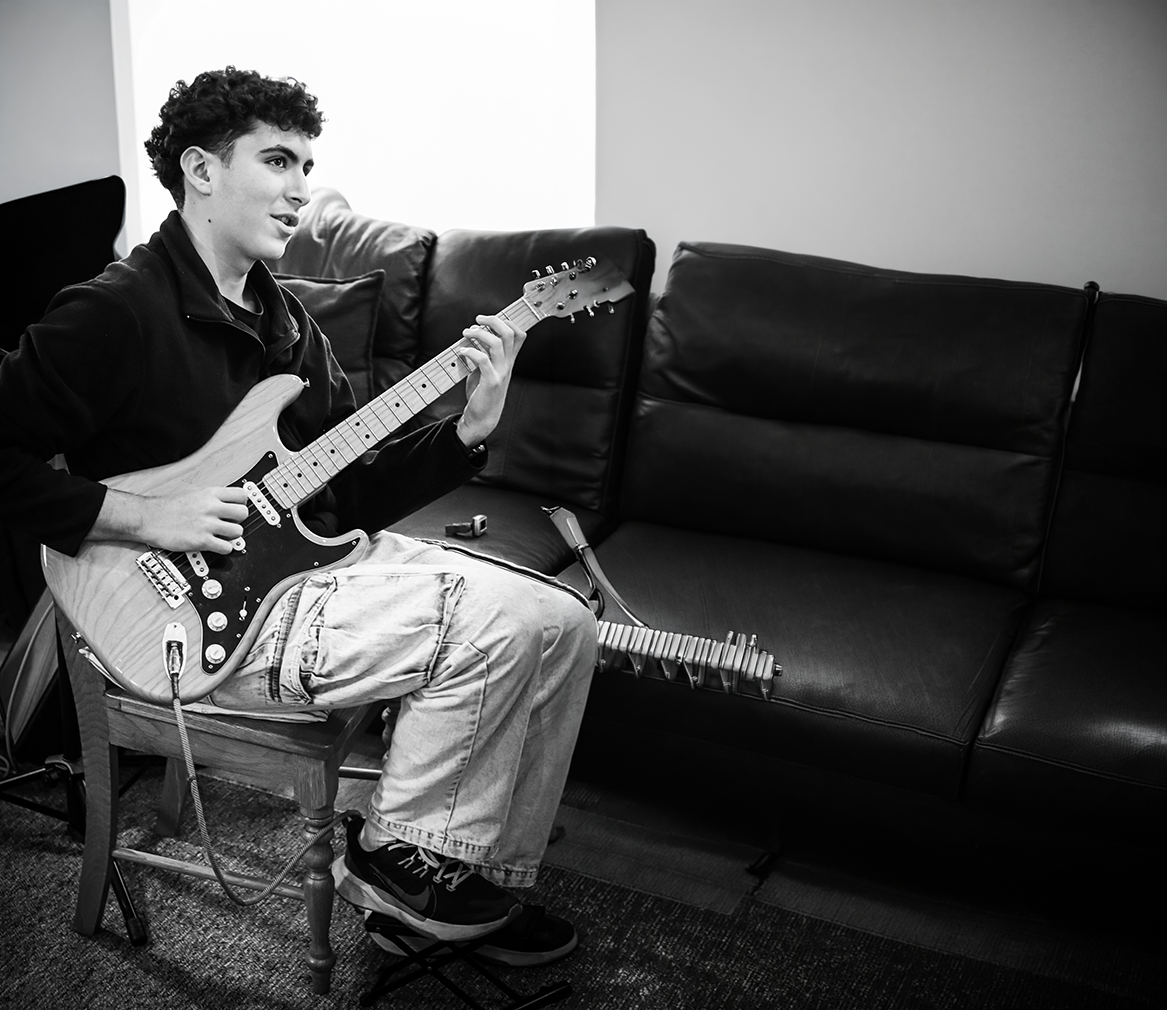 A young man sitting on a wooden chair playing an electric guitar in a room with a black leather couch and a plain wall in the background.