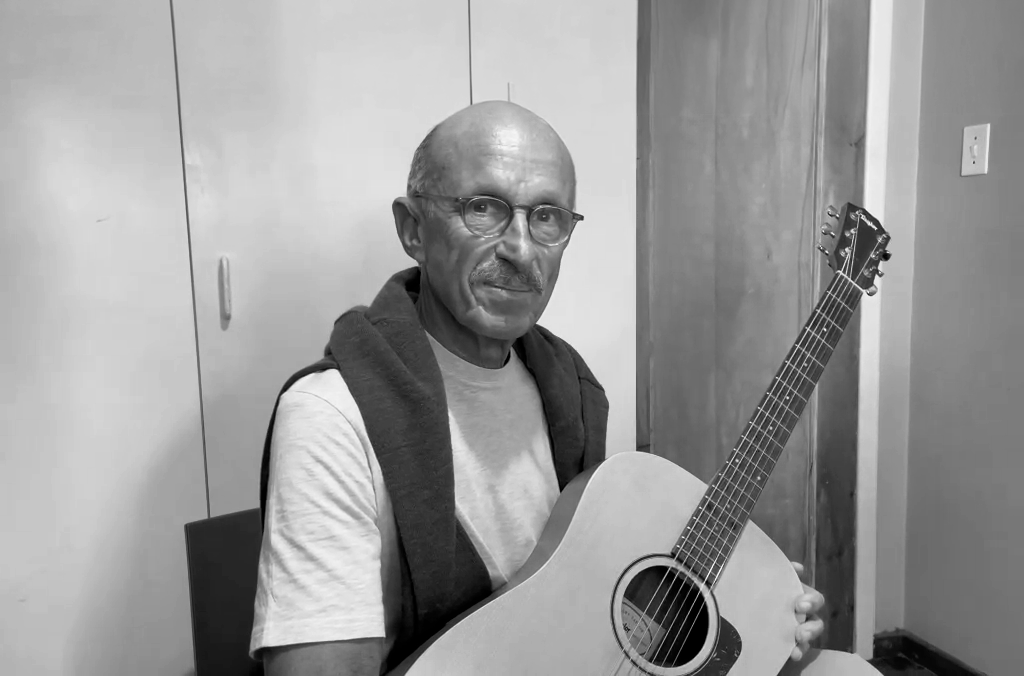 An older man with glasses holding an acoustic guitar in a room with plain walls and a closet door.