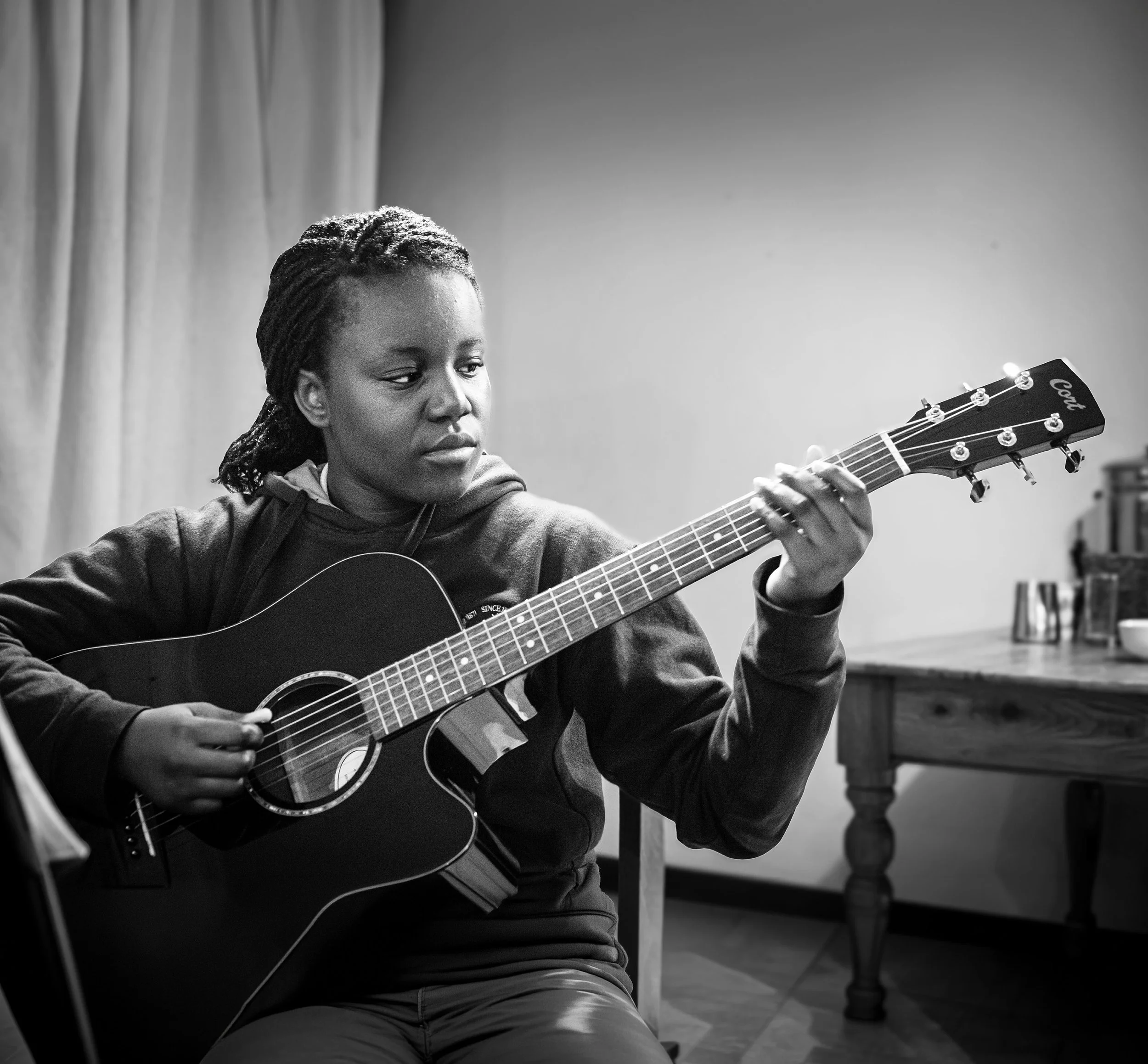 A woman with braided hair playing an acoustic guitar indoors, with a curtain and table in the background.