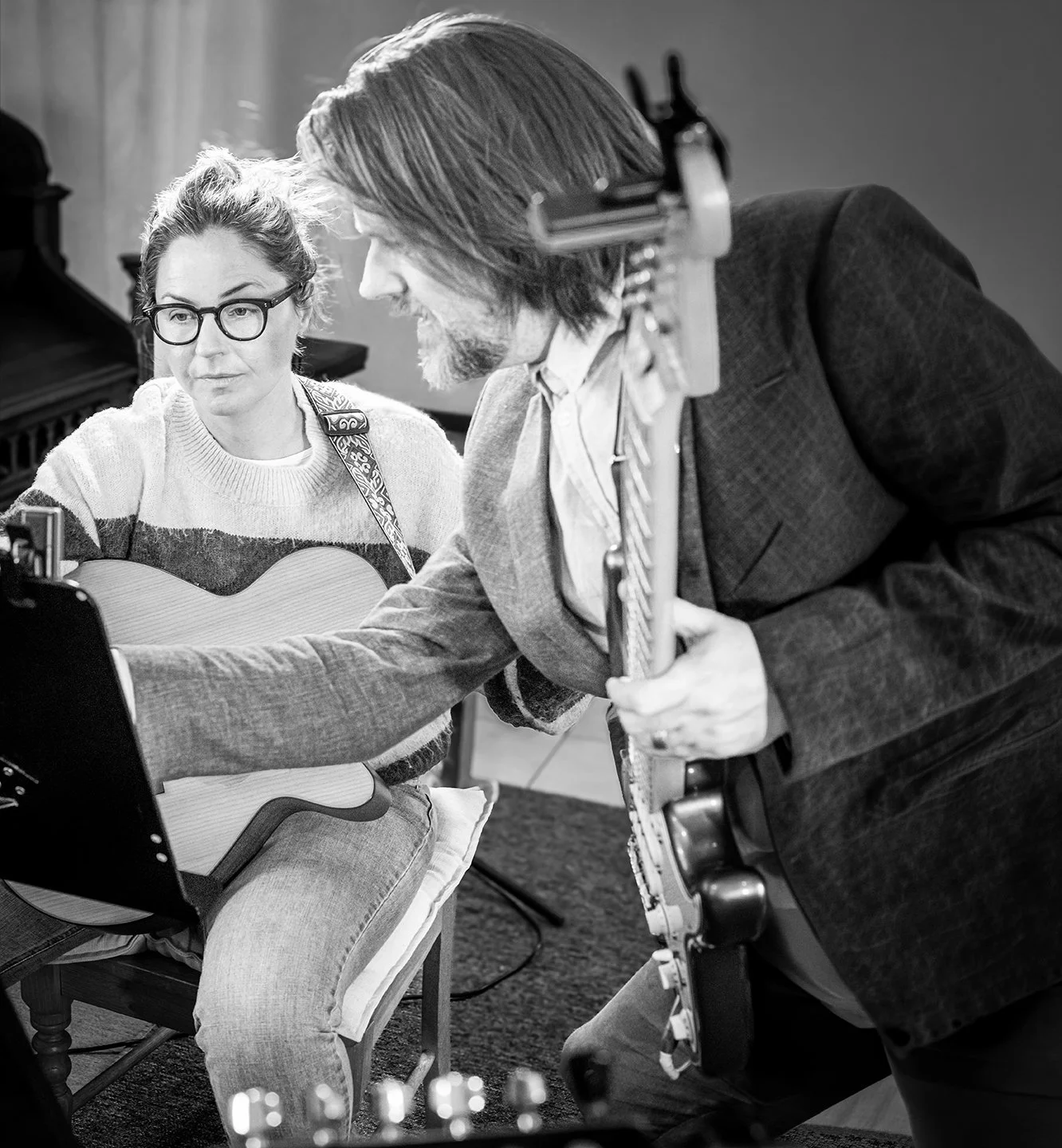 Two musicians practicing; a woman with glasses holding an acoustic guitar, and a man with long hair and beard with a saxophone, looking at a music stand or sheet music.