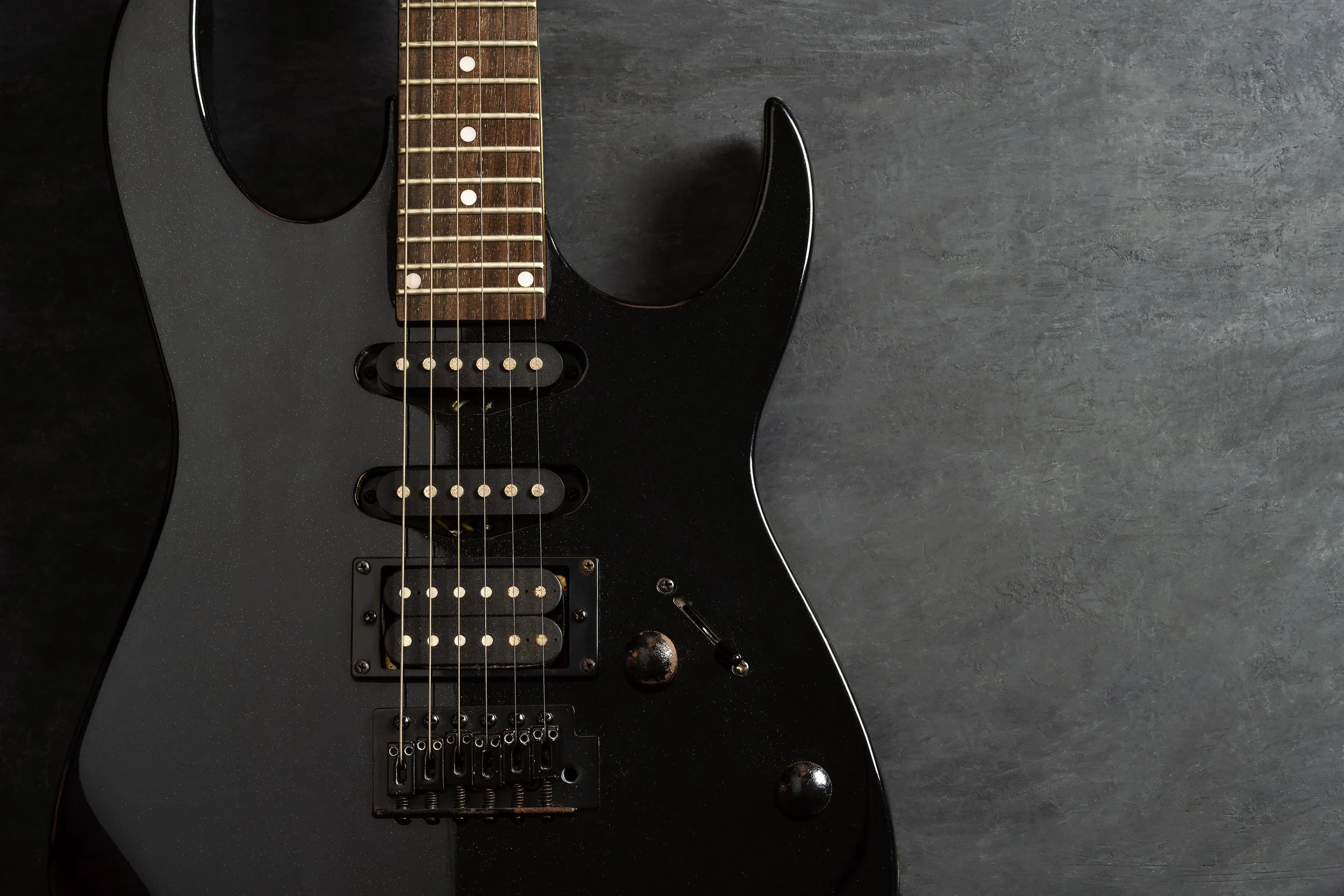 Close-up of a black electric guitar with a dark wooden fingerboard, six strings, and six pickups, lying on a dark gray surface.