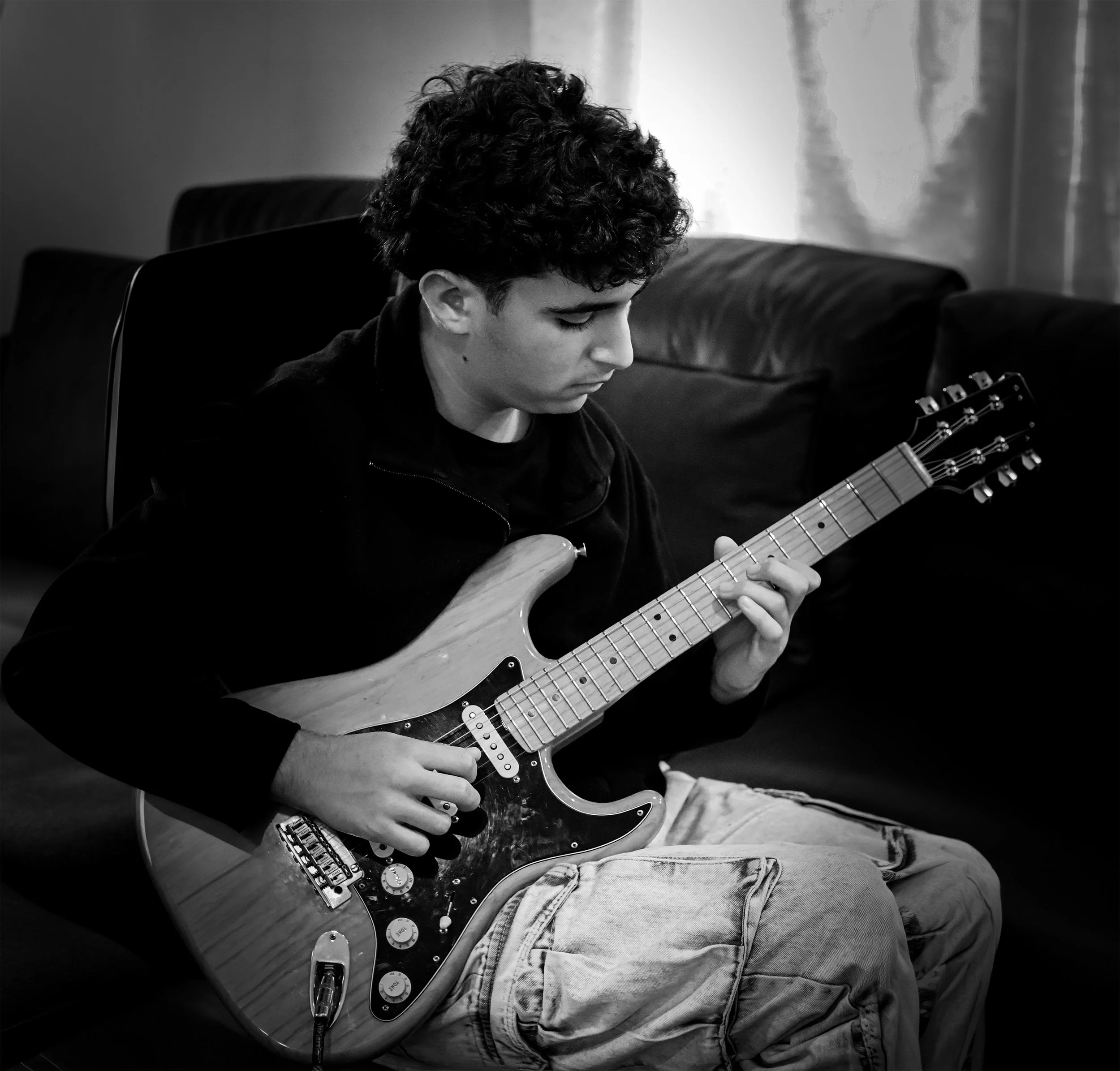 A young man sitting on a couch playing an electric guitar in a room with a window and curtains.