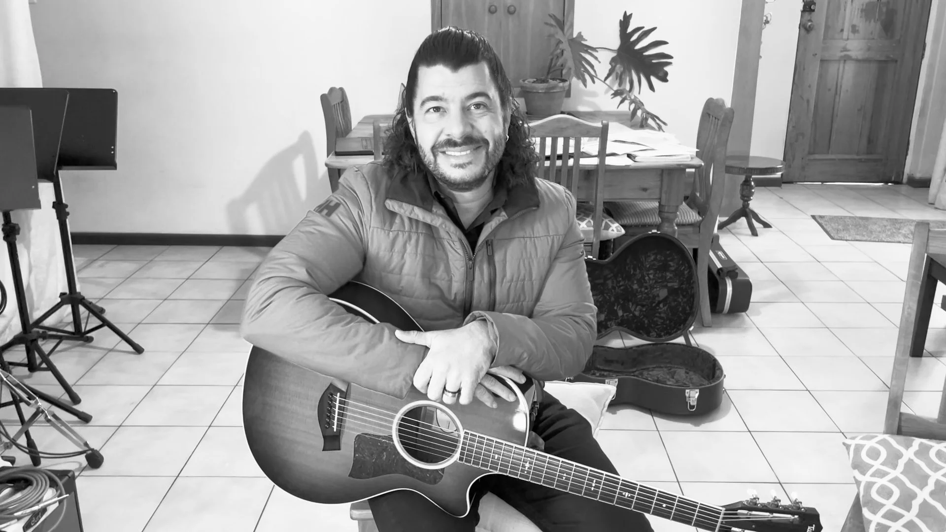 Man with long hair and beard smiling while sitting at a table with an acoustic guitar in a room with a wooden dining table, chairs, potted plants, and music equipment.
