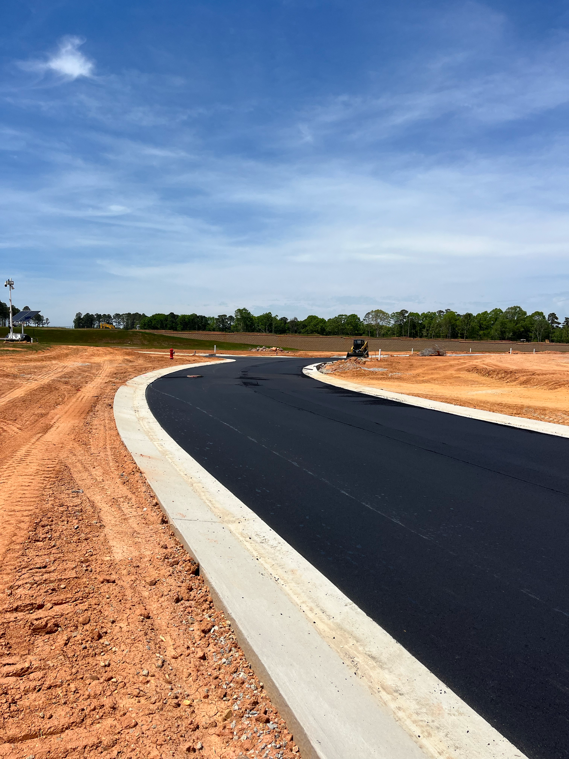 Newly paved black asphalt road with white curb in a rural area under a partly cloudy blue sky, surrounded by orange soil and green trees in the distance.