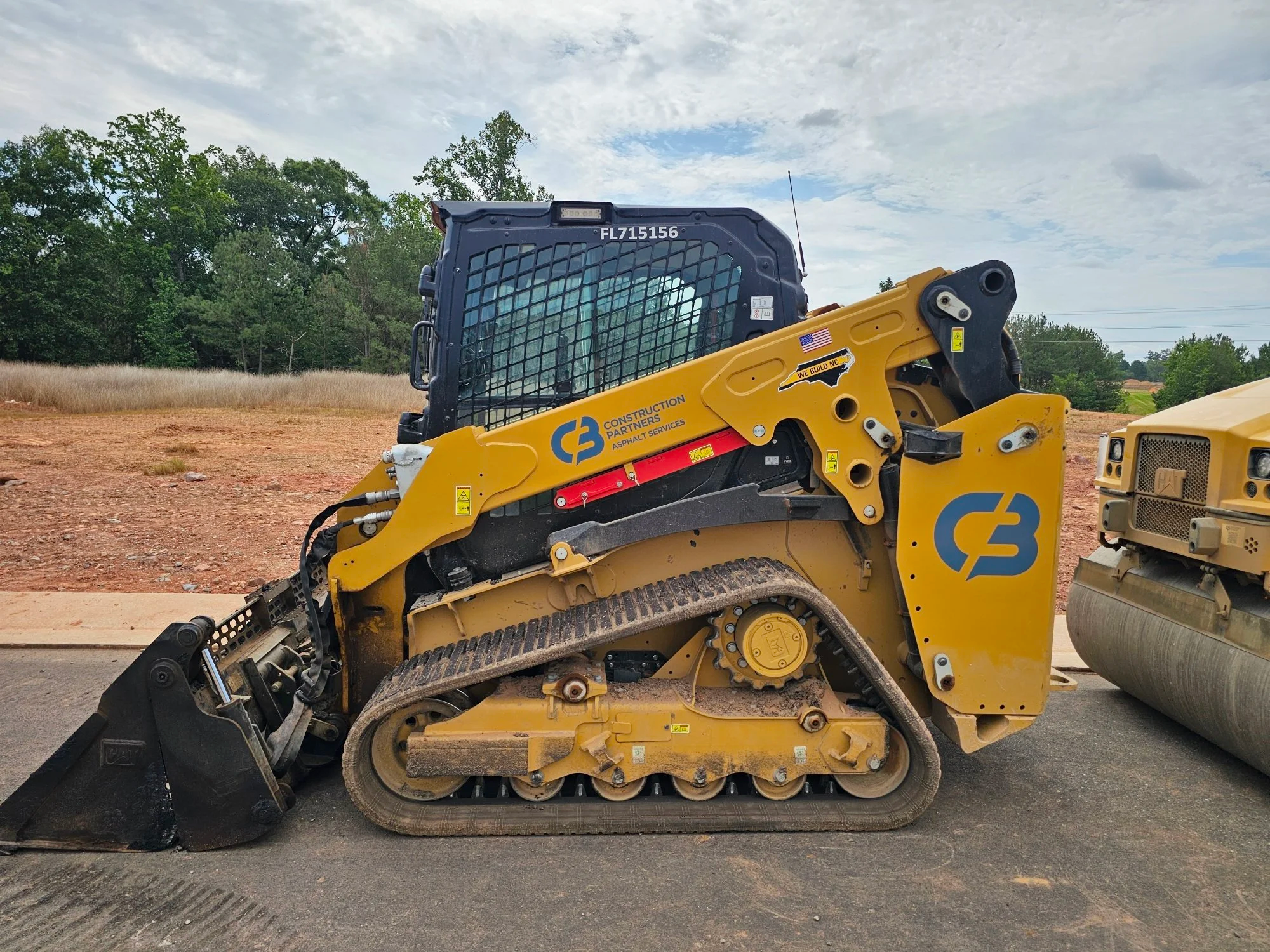 A yellow compact track loader with a black metal mesh cab, parked on a construction site with dirt ground and a backdrop of green trees and cloudy sky, with the C&B Construction Partners logo painted on the side.