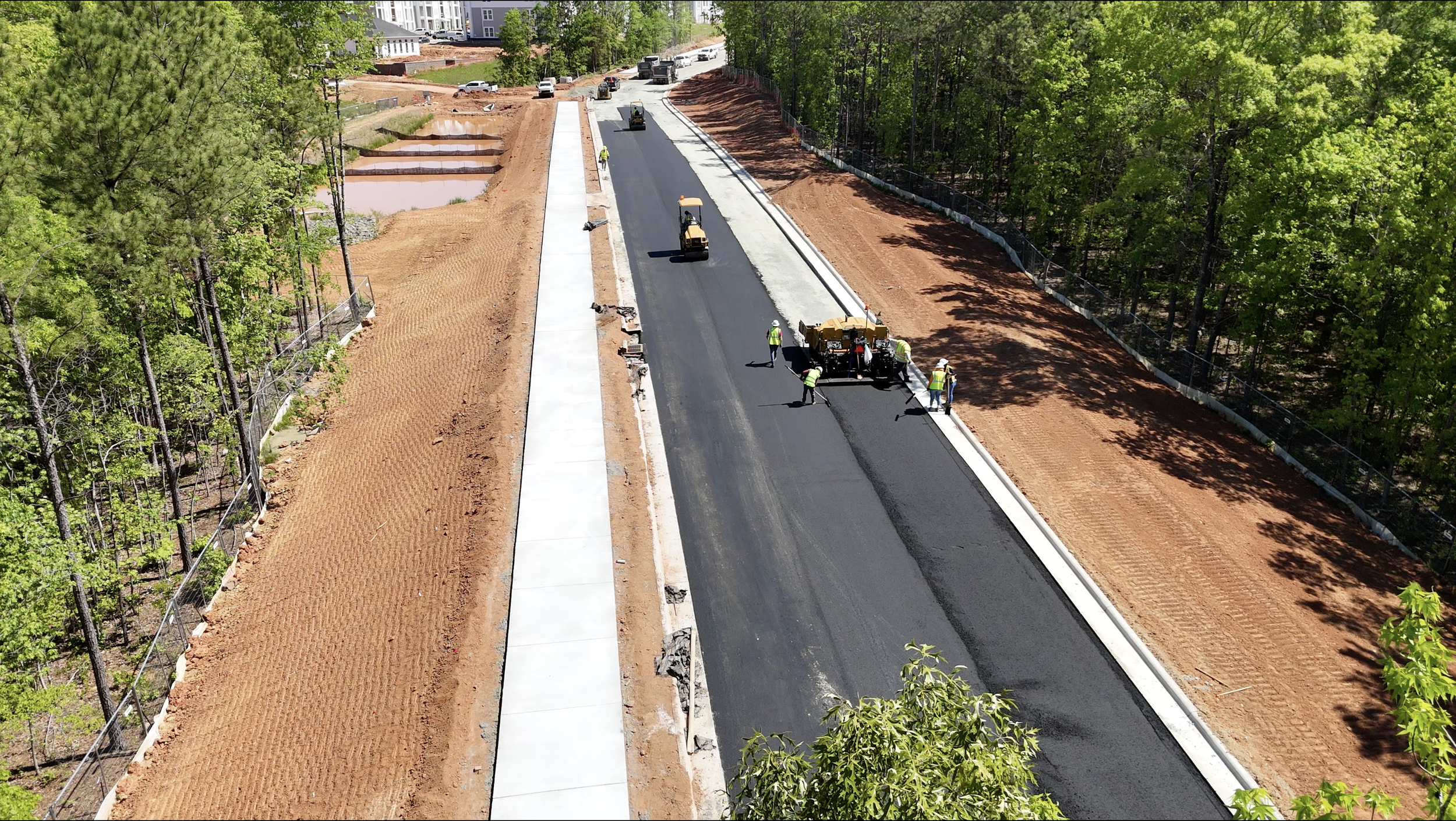 Construction workers are paving a new road along a wooded area, with some machinery and parked vehicles visible in the background.