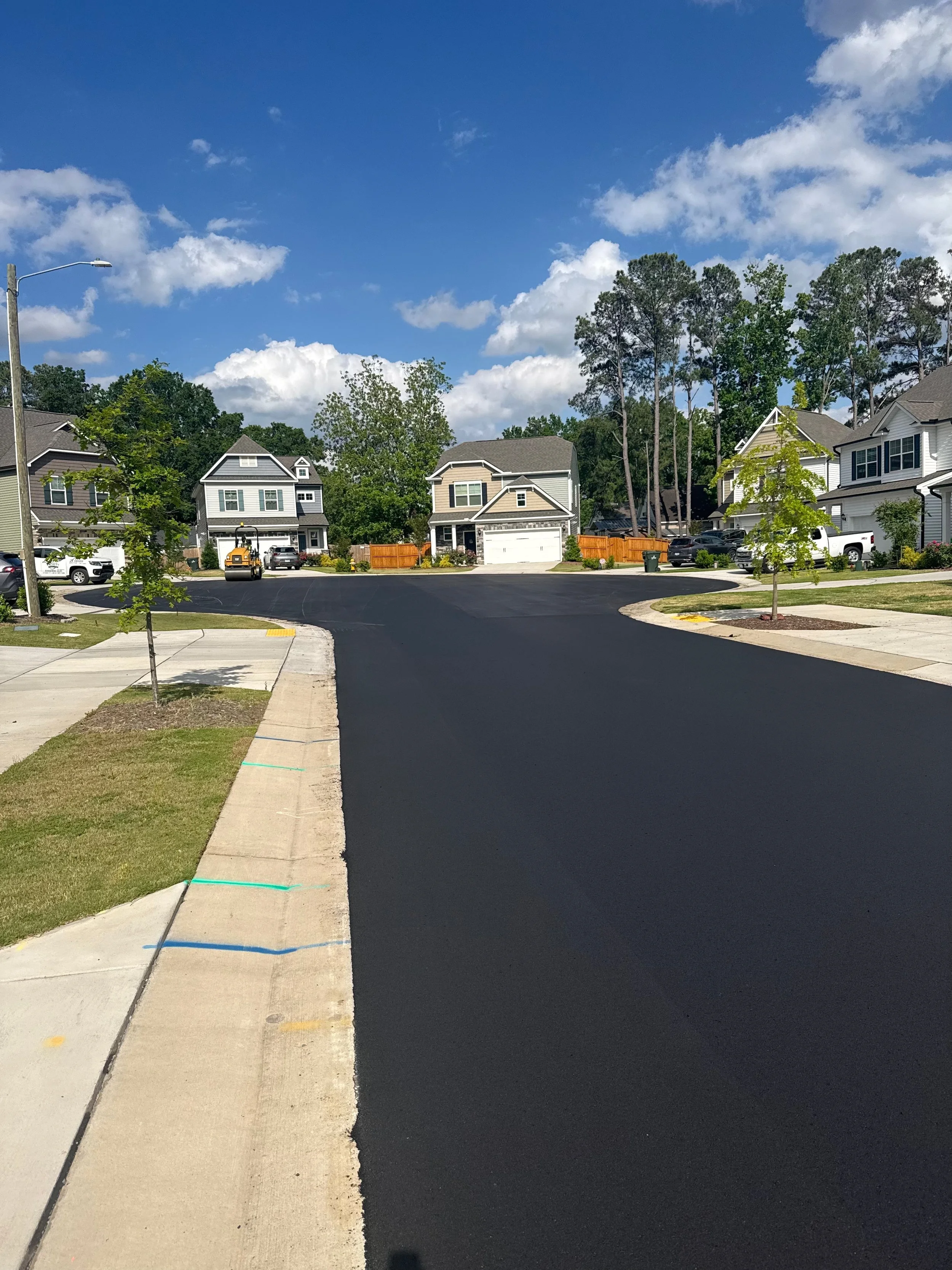 A newly paved black asphalt street in a suburban neighborhood with sidewalk, young trees, and single-family homes under a blue sky with some clouds.