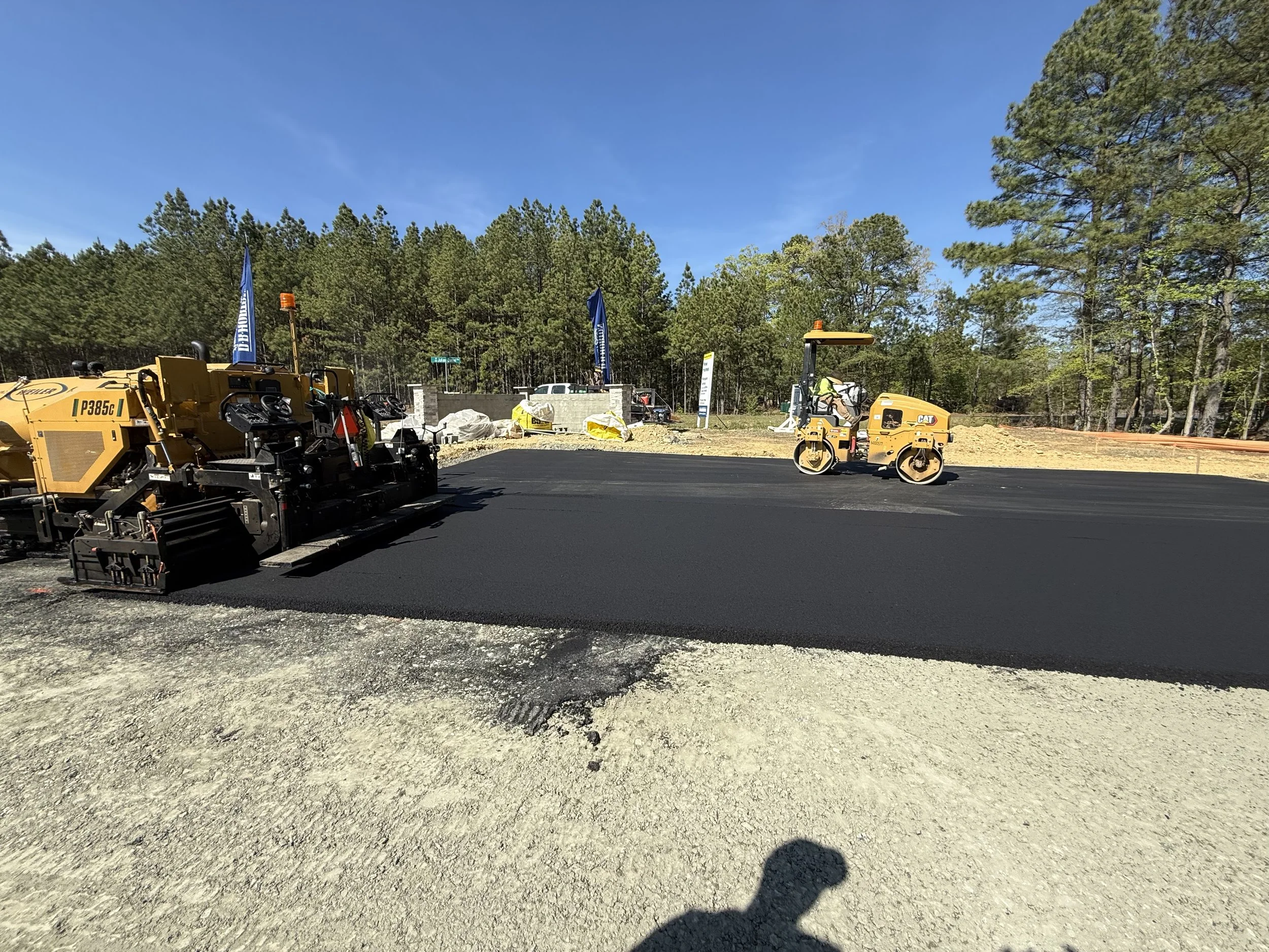 Construction site with asphalt being laid, construction equipment, and a worker operating machinery amidst trees and clear blue sky.