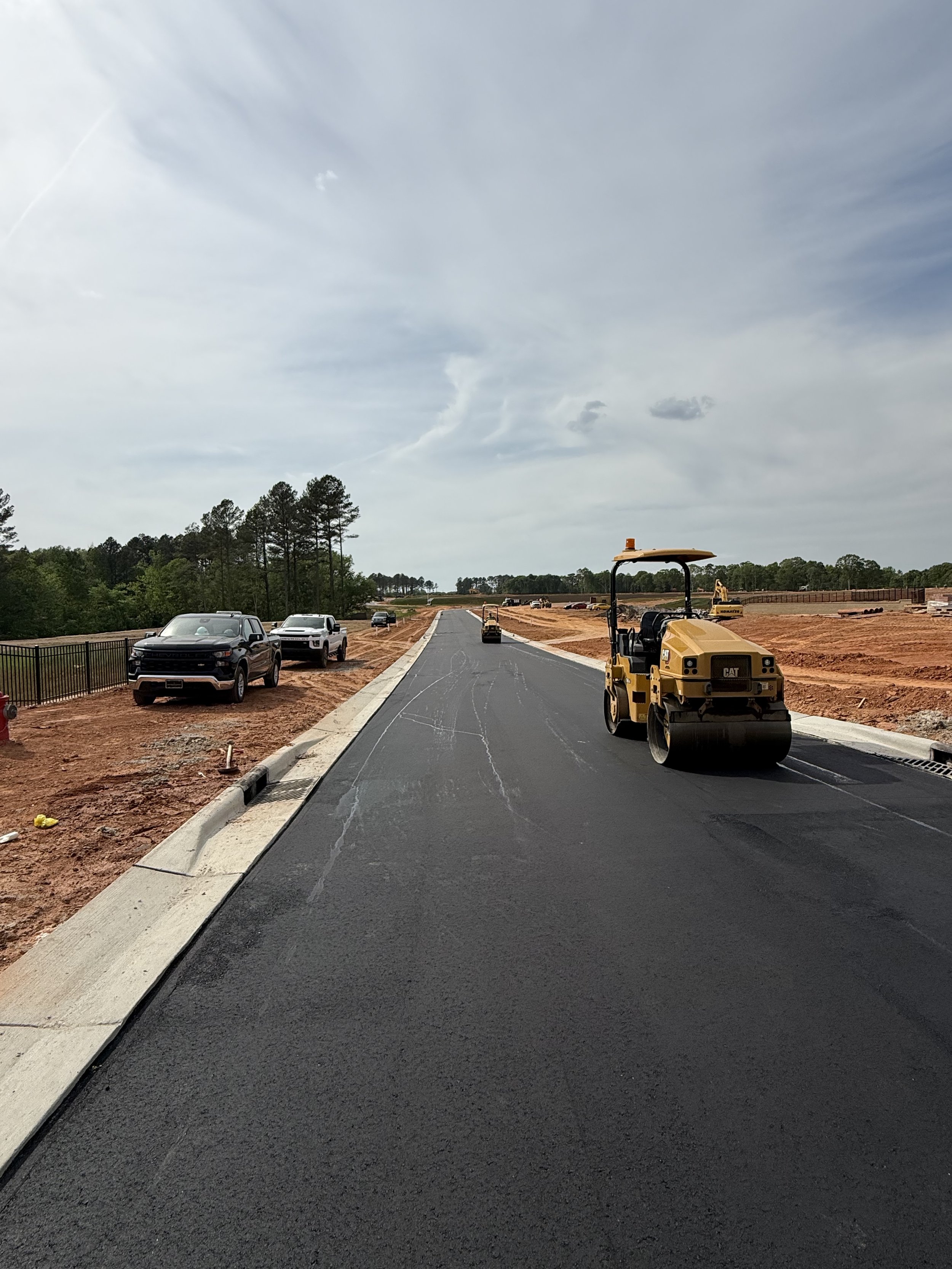 Construction site with freshly paved road, construction vehicles including a roller and excavator, parked cars along the sides, and a cloudy sky overhead.