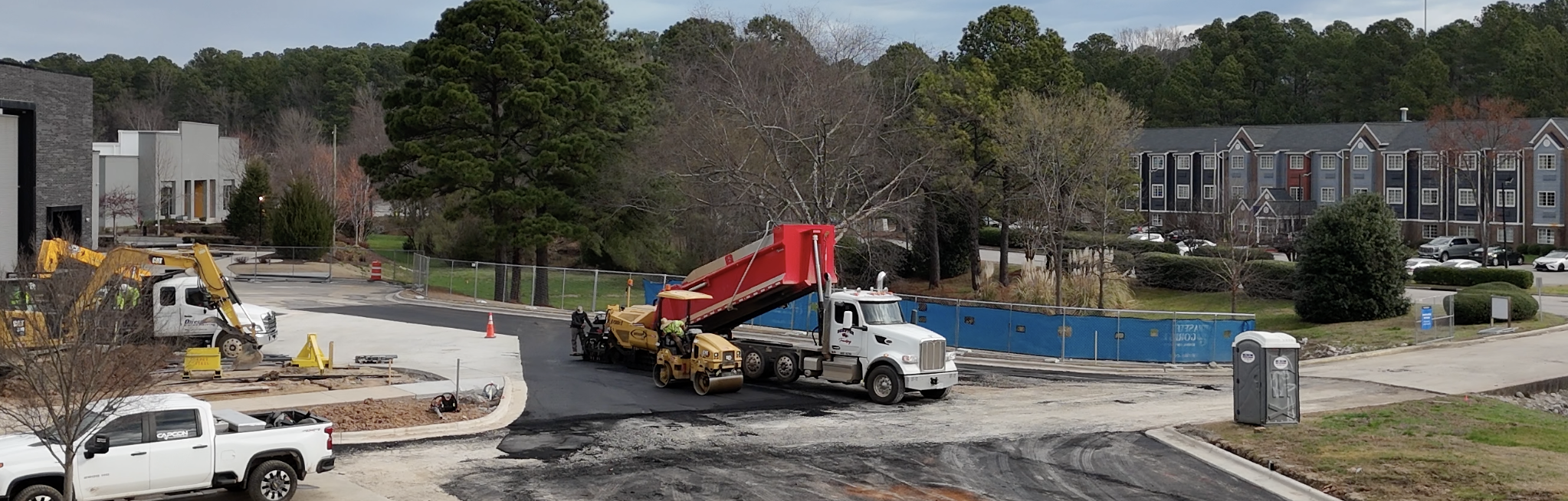 Construction site with a dump truck and worker, surrounded by trees and residential buildings in the background.