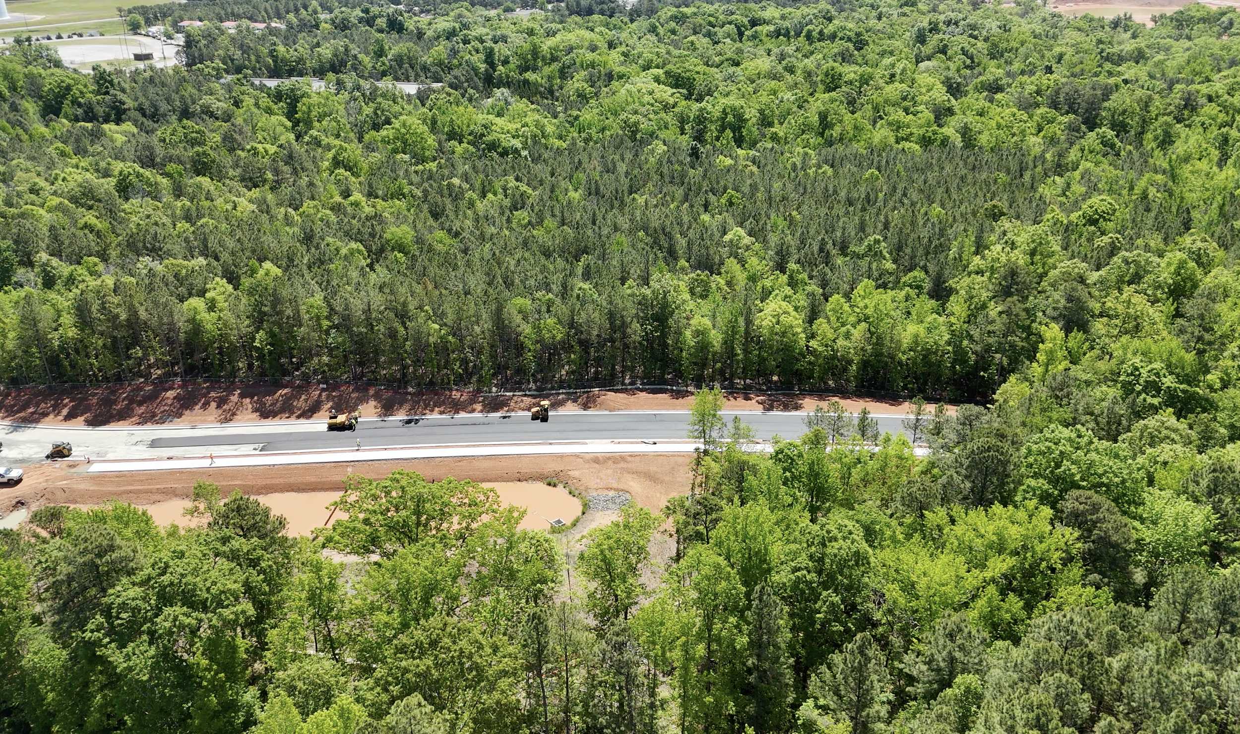 An aerial view of a road under construction amidst a green forest. Several construction vehicles are working on the road, with trees and dense forest surrounding the area.