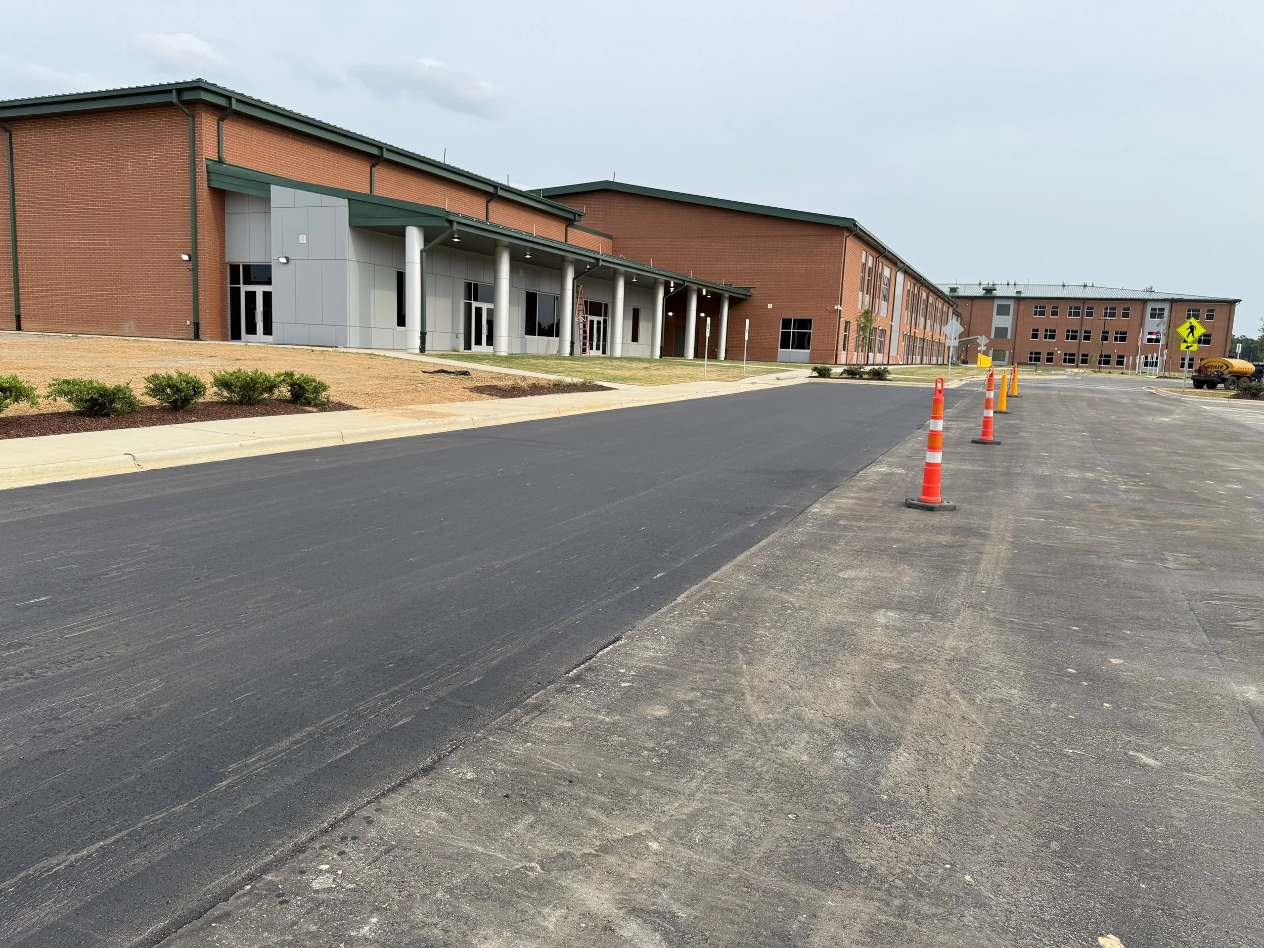 An empty parking lot with recently repaved asphalt, orange traffic cones, a brick and gray building with glass doors and windows, and a cloudy sky.