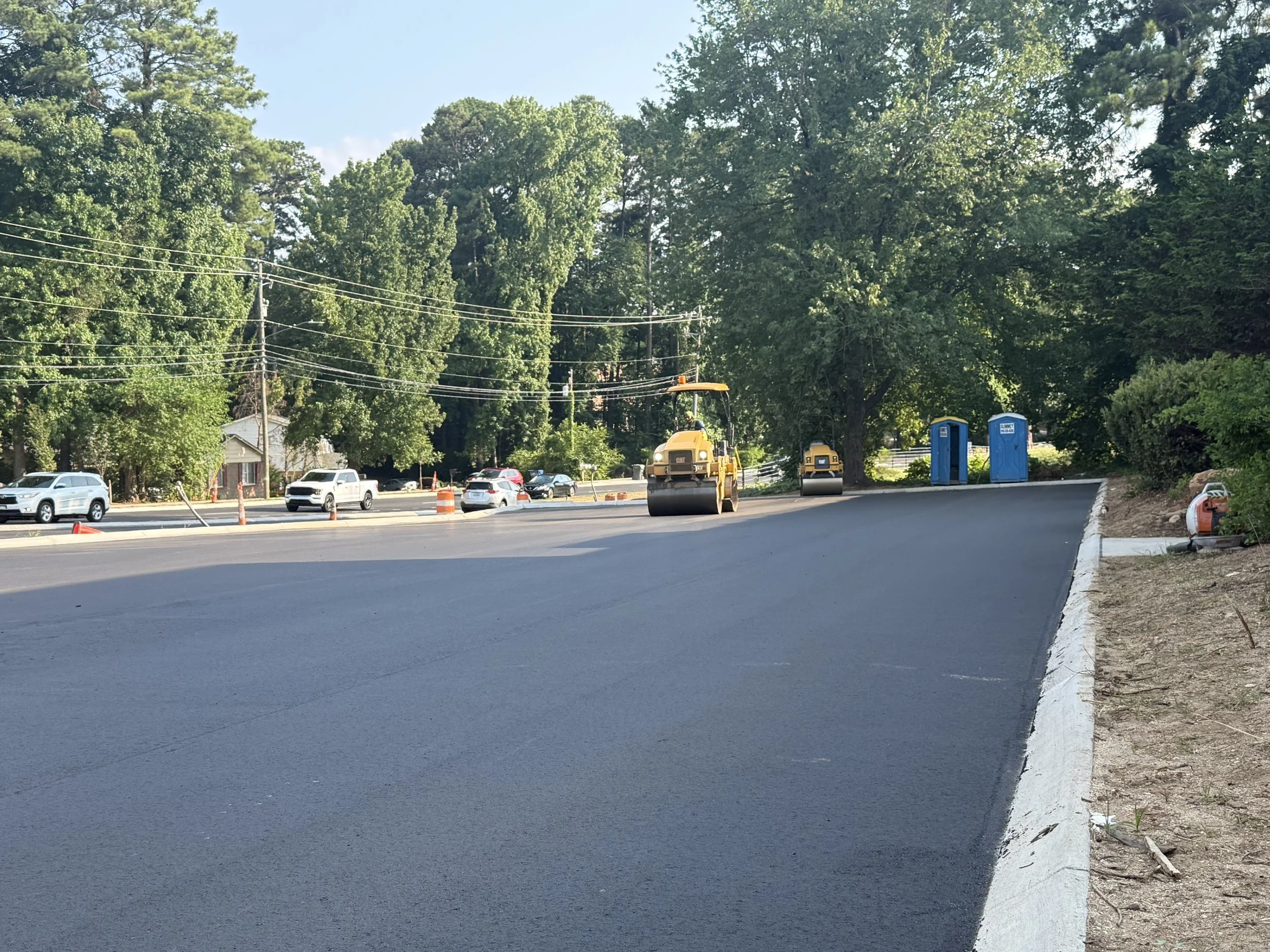 Construction crew working on road repair with asphalt rollers, parked cars, trees, portable toilets, and traffic cones.