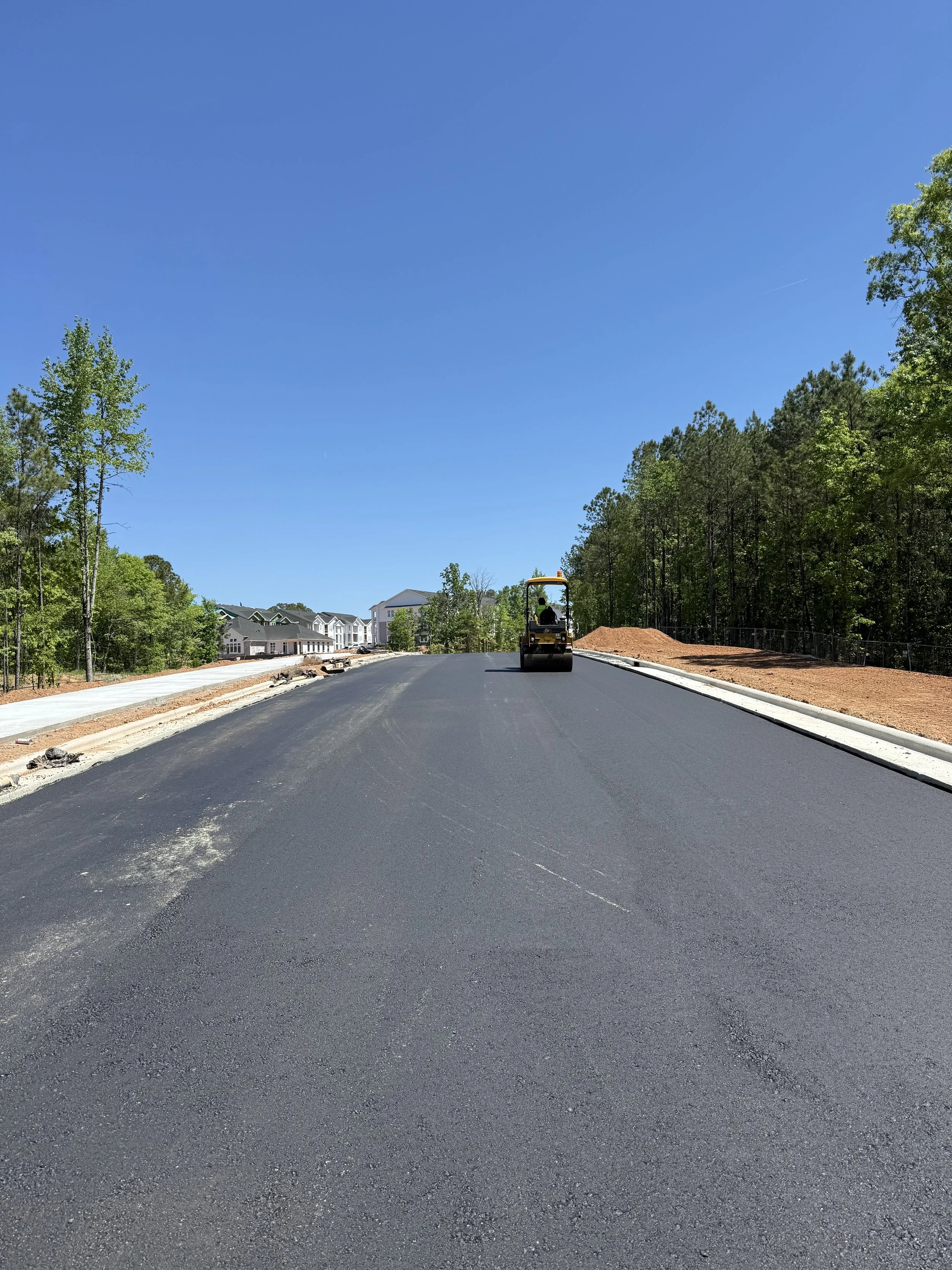 A road construction scene with a black asphalt roller compactor in the center, working on freshly paved black asphalt. To the left, there are some trees and houses in the background. To the right, there is a dirt area with a chain-link fence and surr