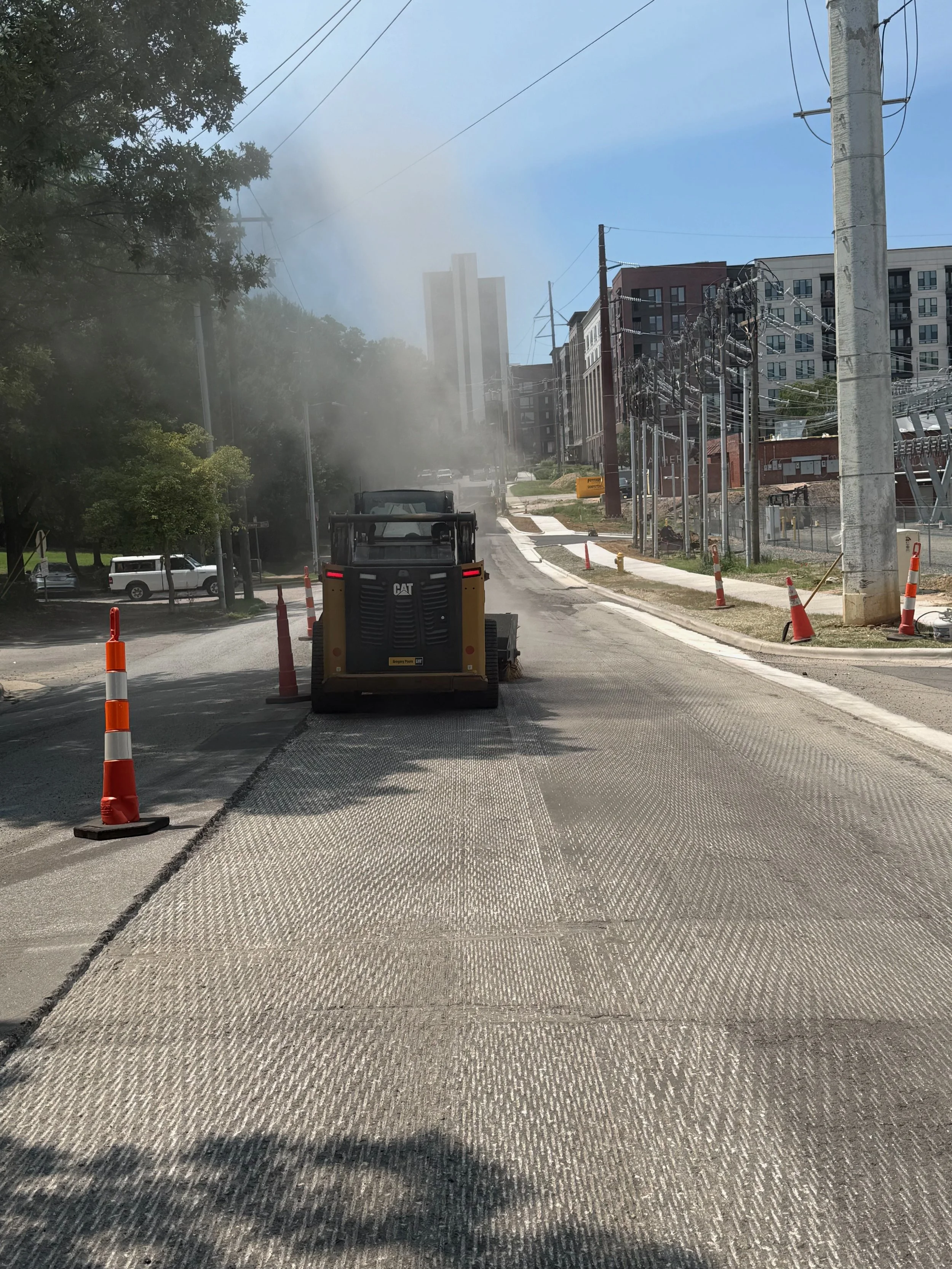 Construction site with a construction vehicle in the foreground, orange traffic cones, and snow or debris on the street. Buildings and utility poles in the background, with some steam or dust rising from the area.