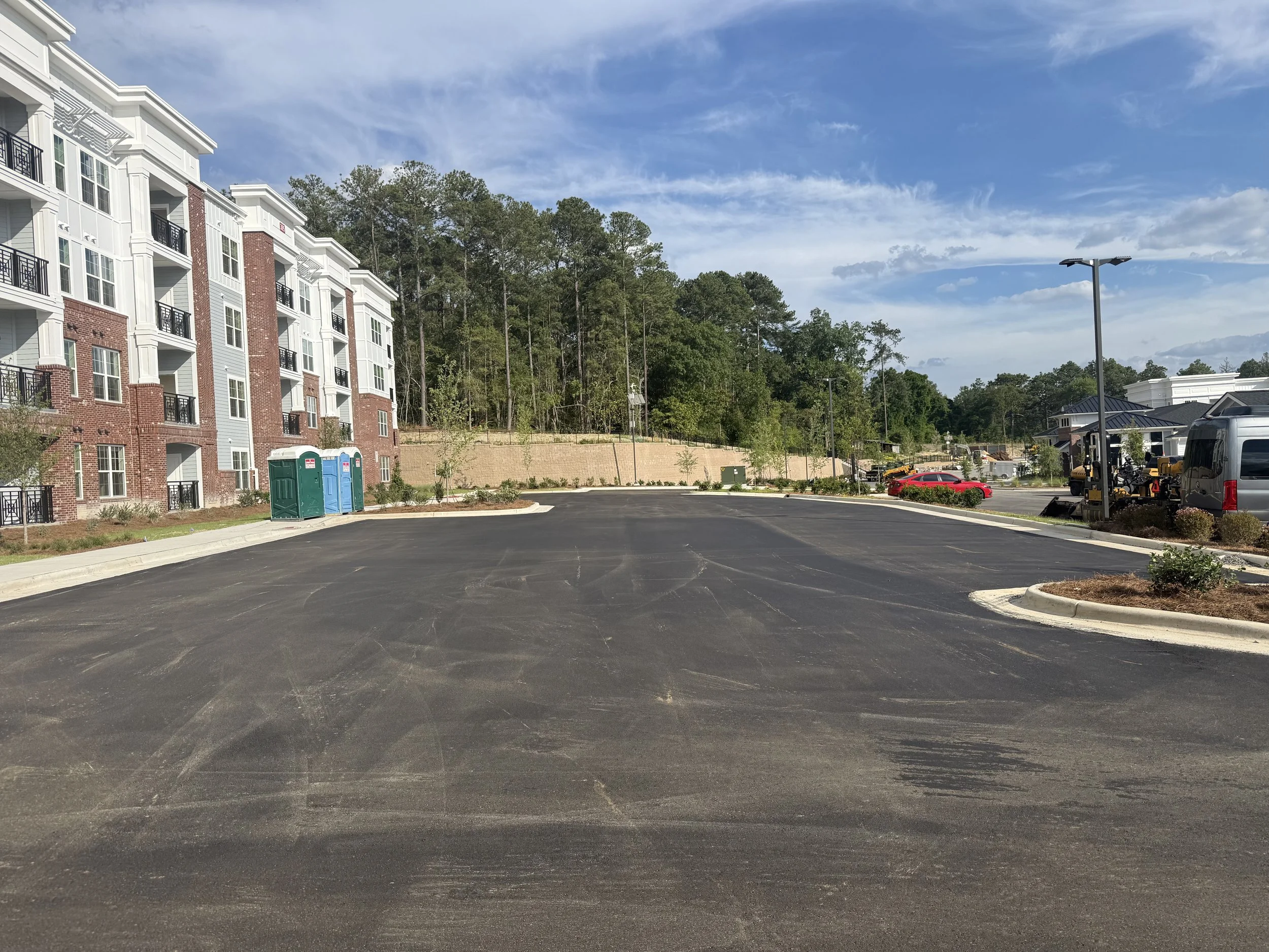 A parking lot in front of an apartment complex with trees and construction equipment in the background, sunny sky with some clouds.