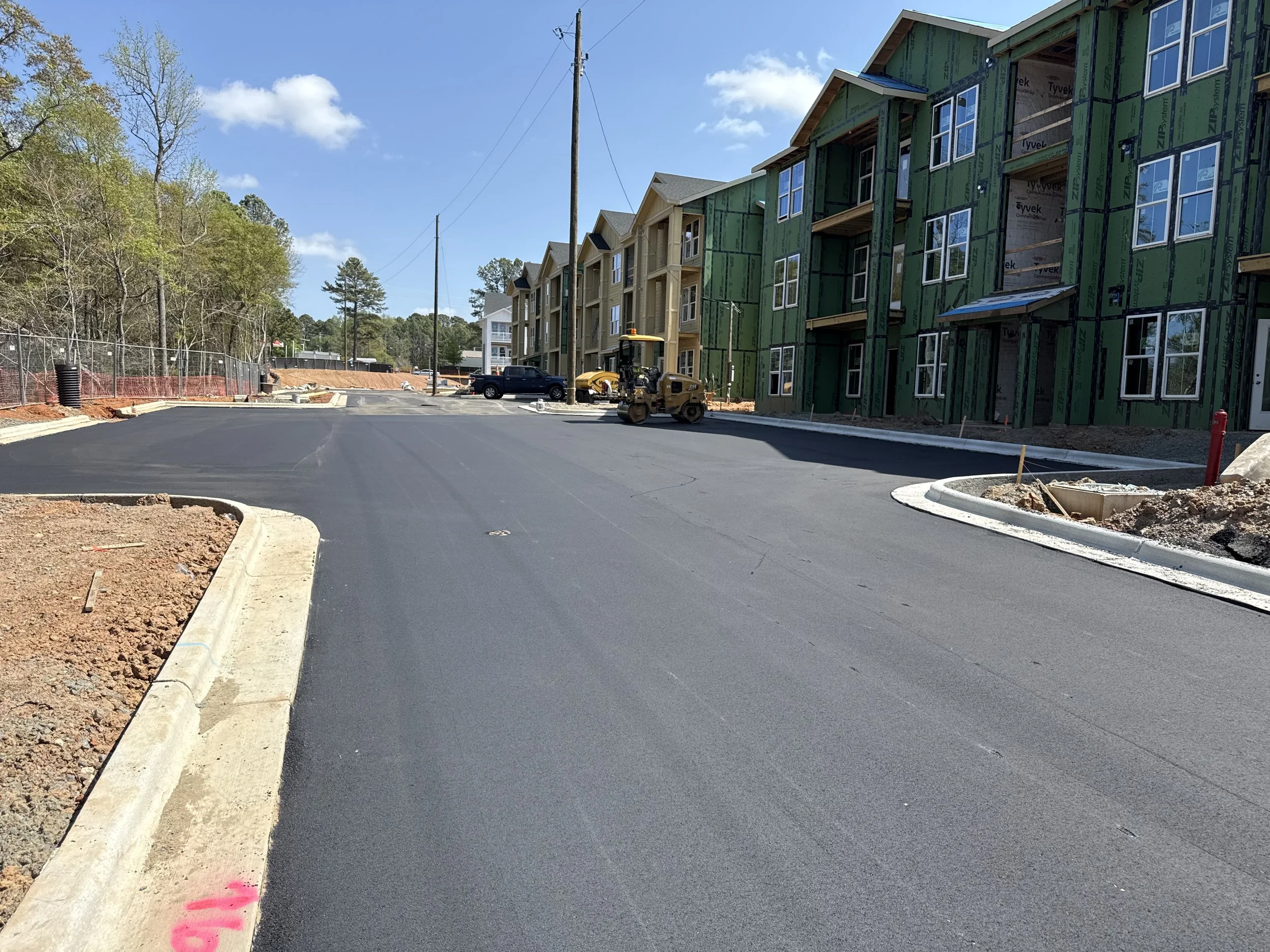 Construction site with newly paved asphalt road, partially constructed apartment buildings with green sheathing, a small roller compactor, parked trucks, a chain-link fence on the left, and a utility pole with wires, under a partly cloudy sky.