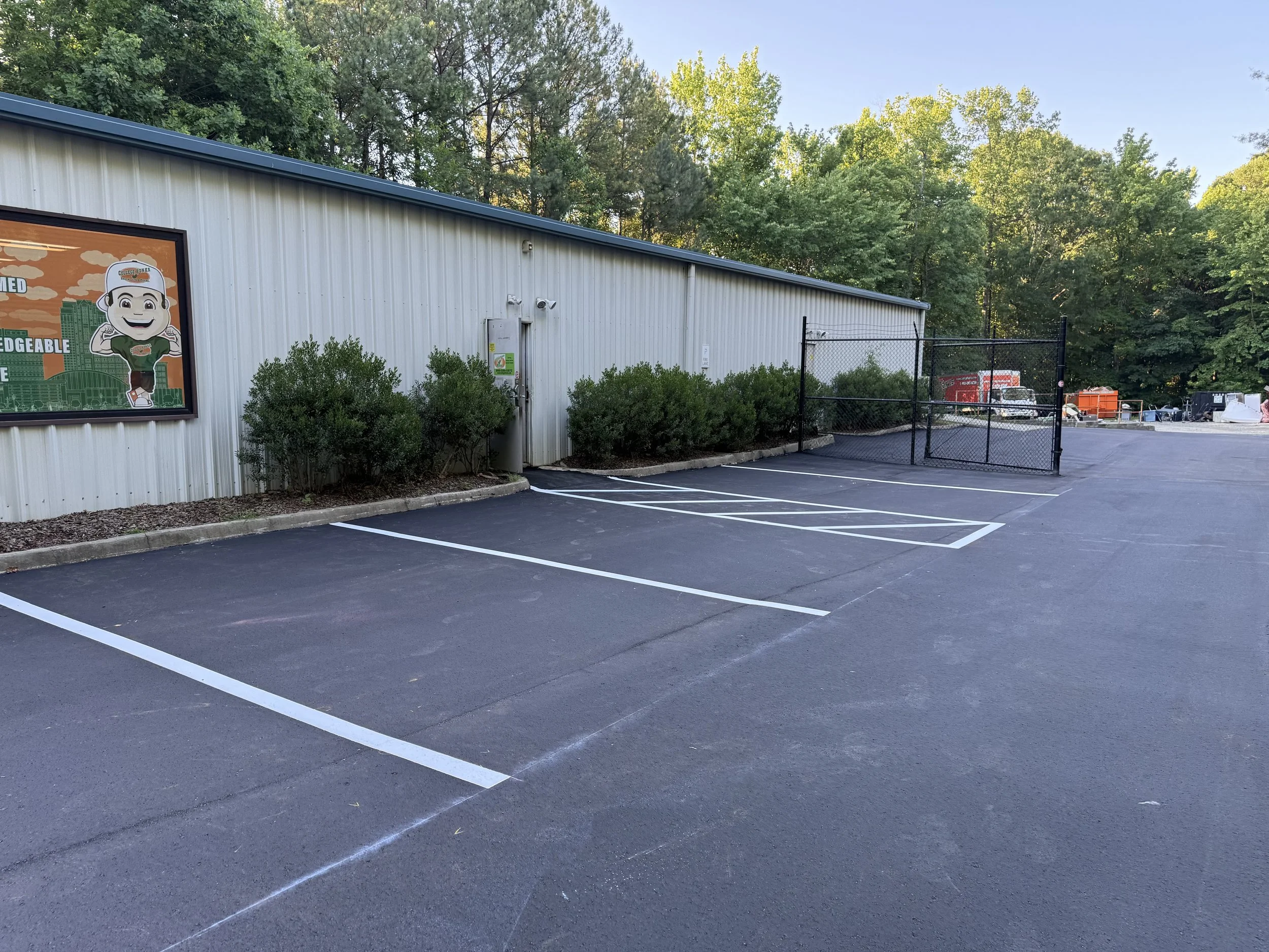 Empty parking lot with striped white parking lines, a white building in the background, surrounded by green trees under a clear blue sky.