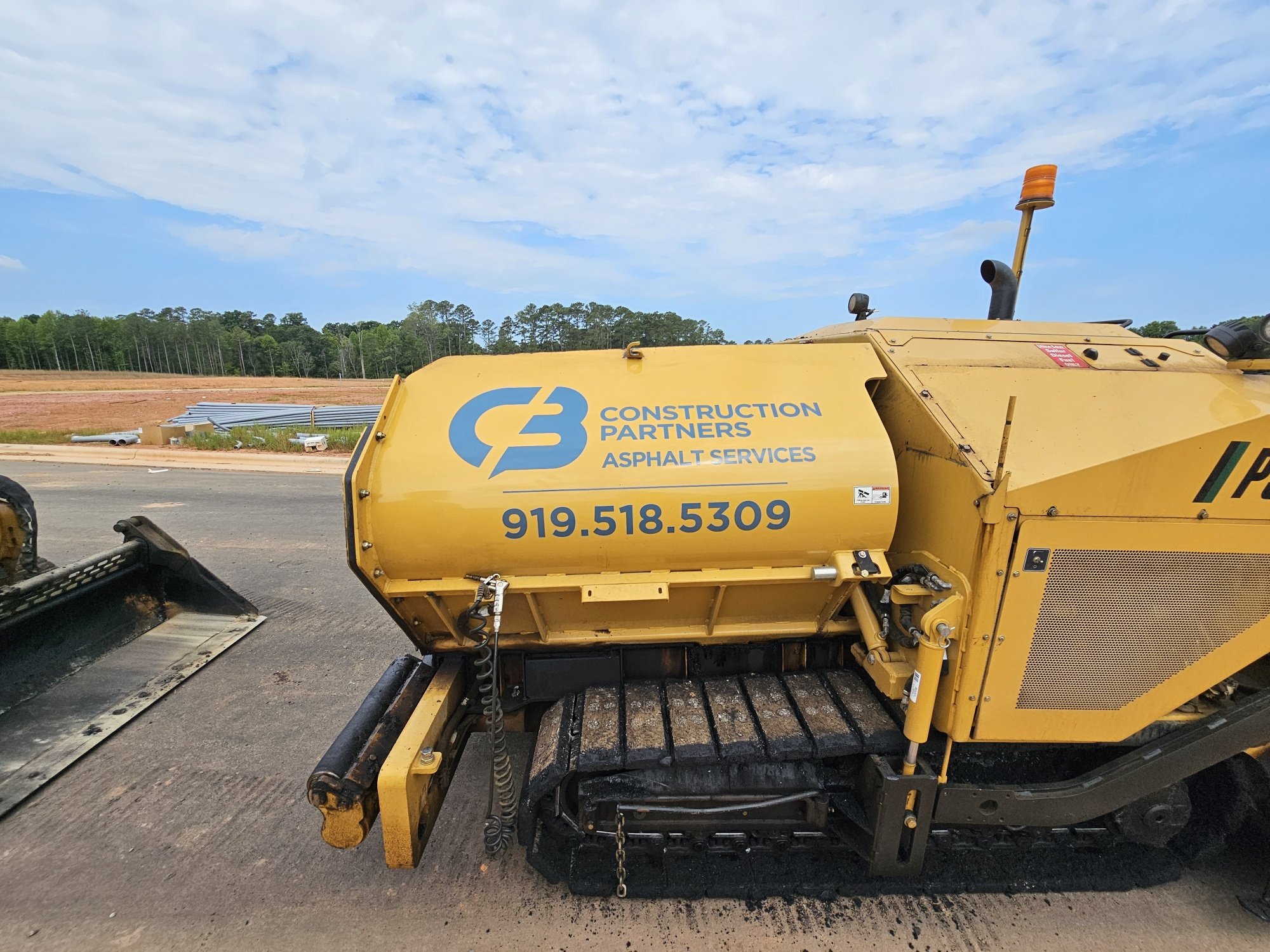 Close-up of yellow road construction machine with the logo and contact info for C&B Construction Partners, on a construction site with soil and trees in the background.