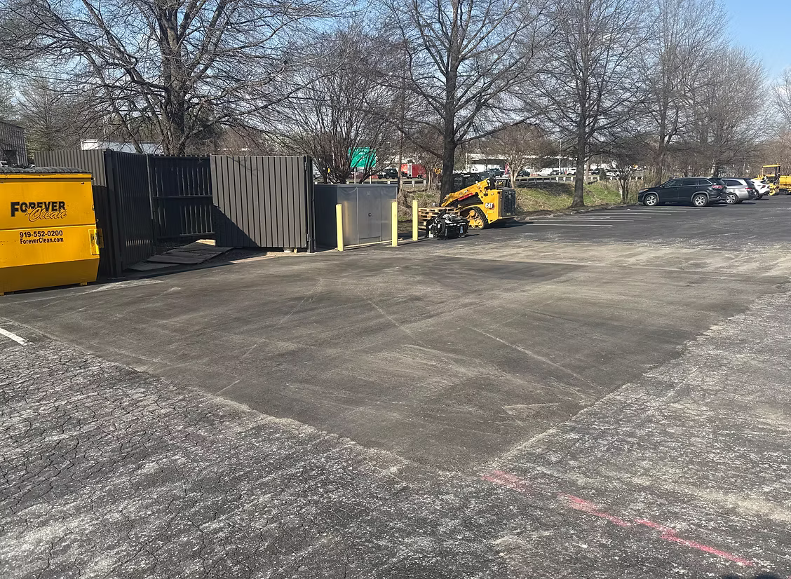 A recently patched section of pavement, in a parking lot parking lot with trees, a yellow dumpster, and construction equipment in the background.