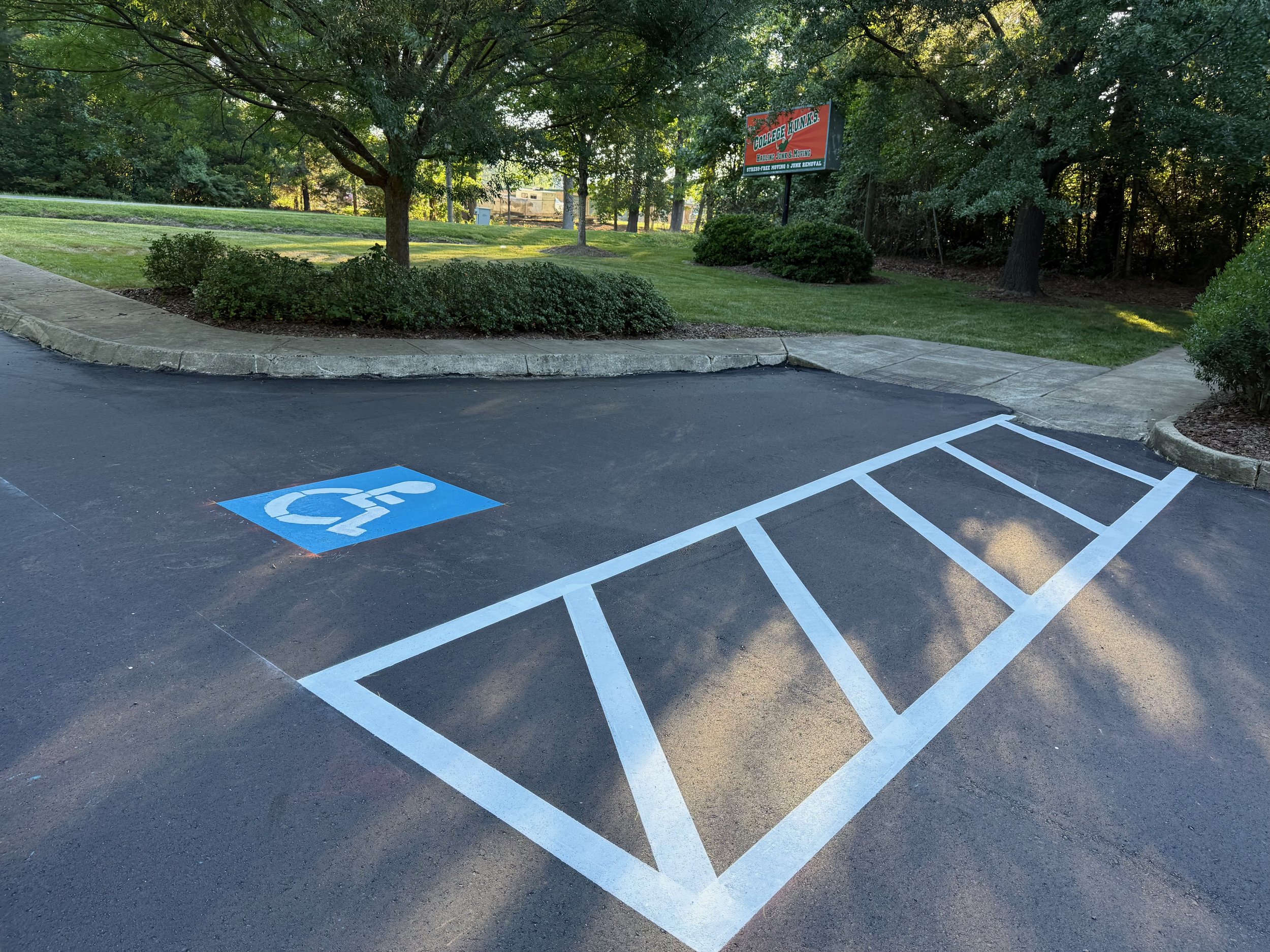 A parking space designated for handicapped with a blue symbol painted on the asphalt. The parking lot is adjacent to a sidewalk, and there are green bushes and trees in the background.