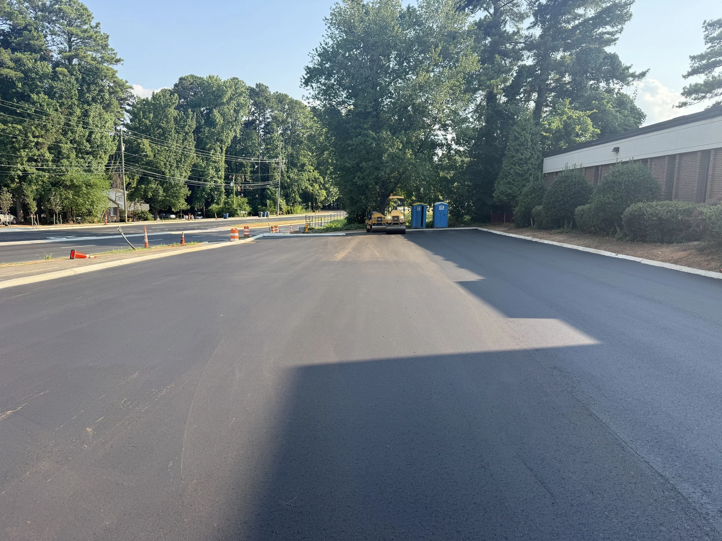 Construction zone with freshly paved road, orange traffic cones, and construction equipment such as a steamroller. There are trees, power lines, and a building on the right side under a clear sky.