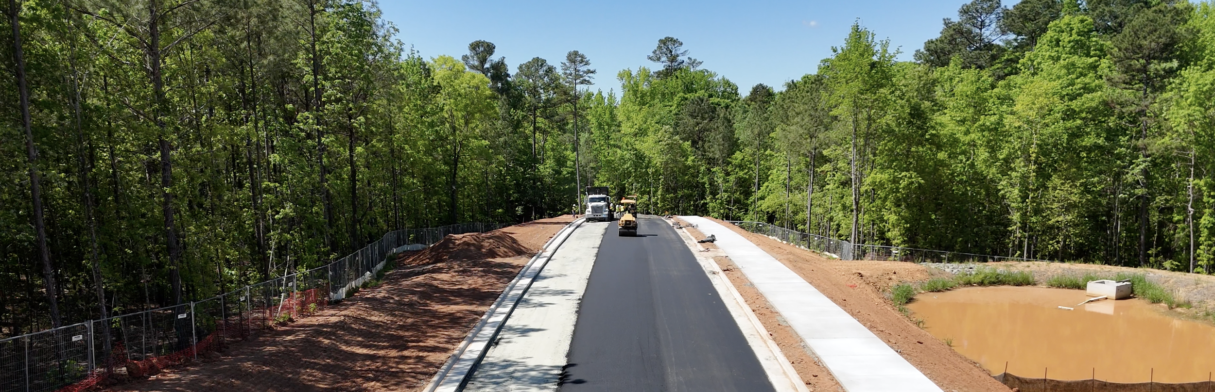 Road construction site with a partly paved road, construction vehicles, and a pond on the right side, surrounded by green trees under a clear blue sky.
