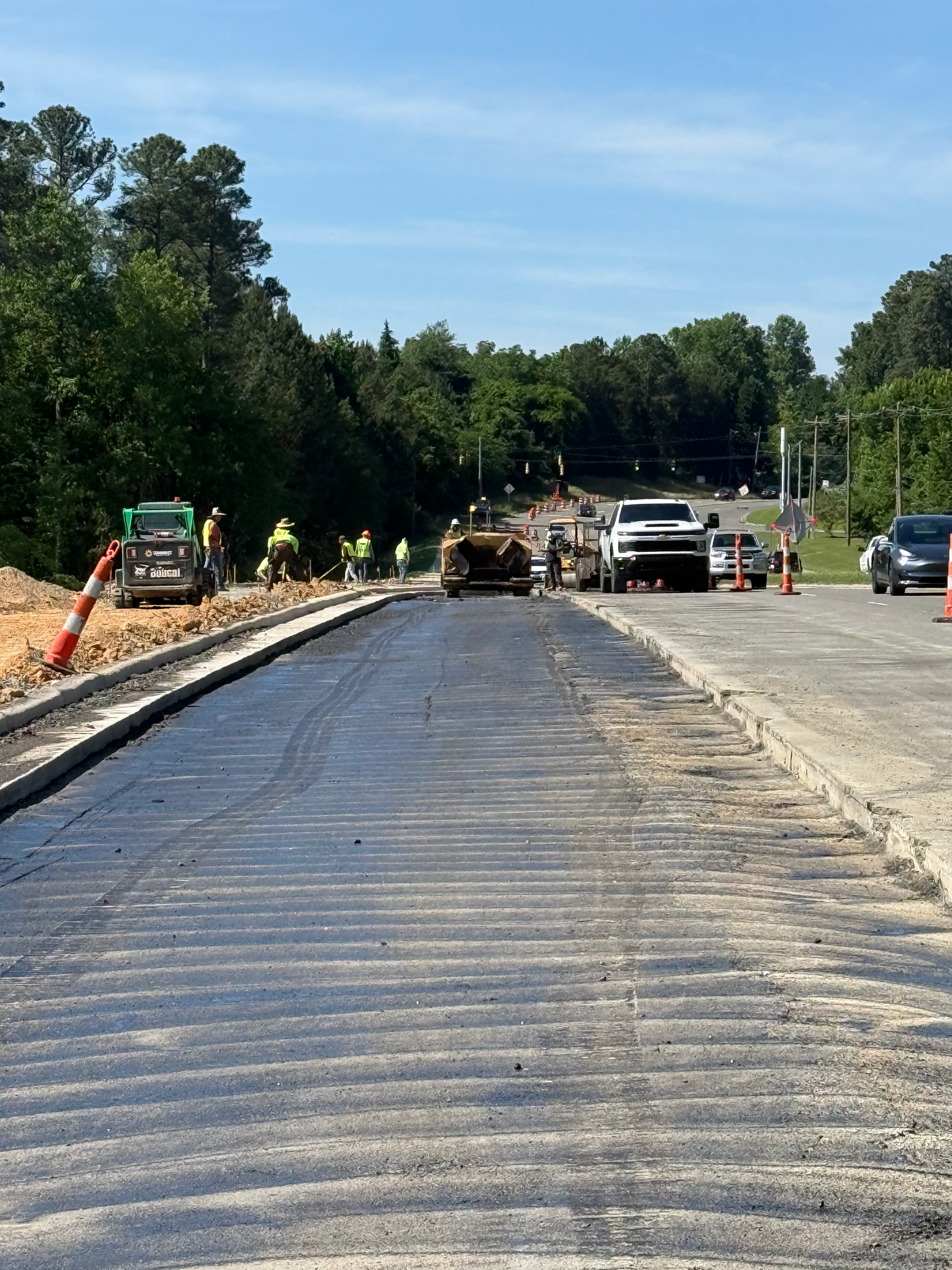 Construction workers and machinery working on paving a road amidst traffic cones and vehicles in a forested area.
