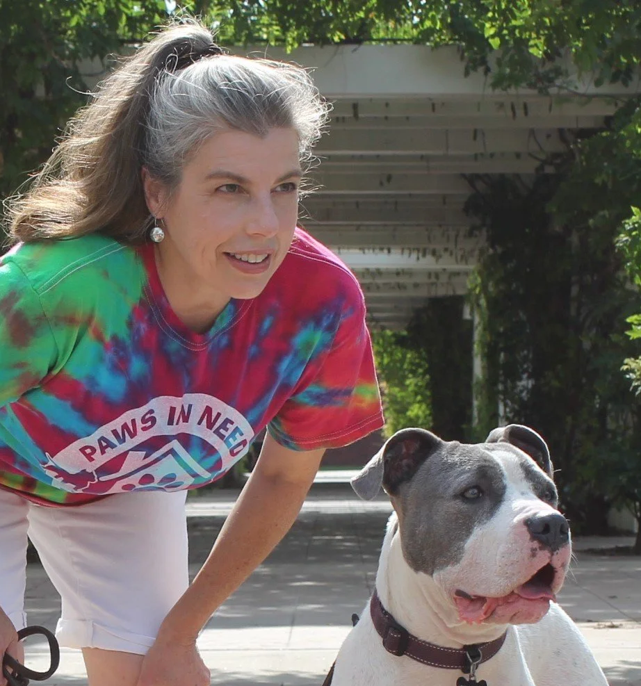 A woman in a colorful tie-dye shirt crouches next to a white and gray pit bull dog outdoors on a sunny day.